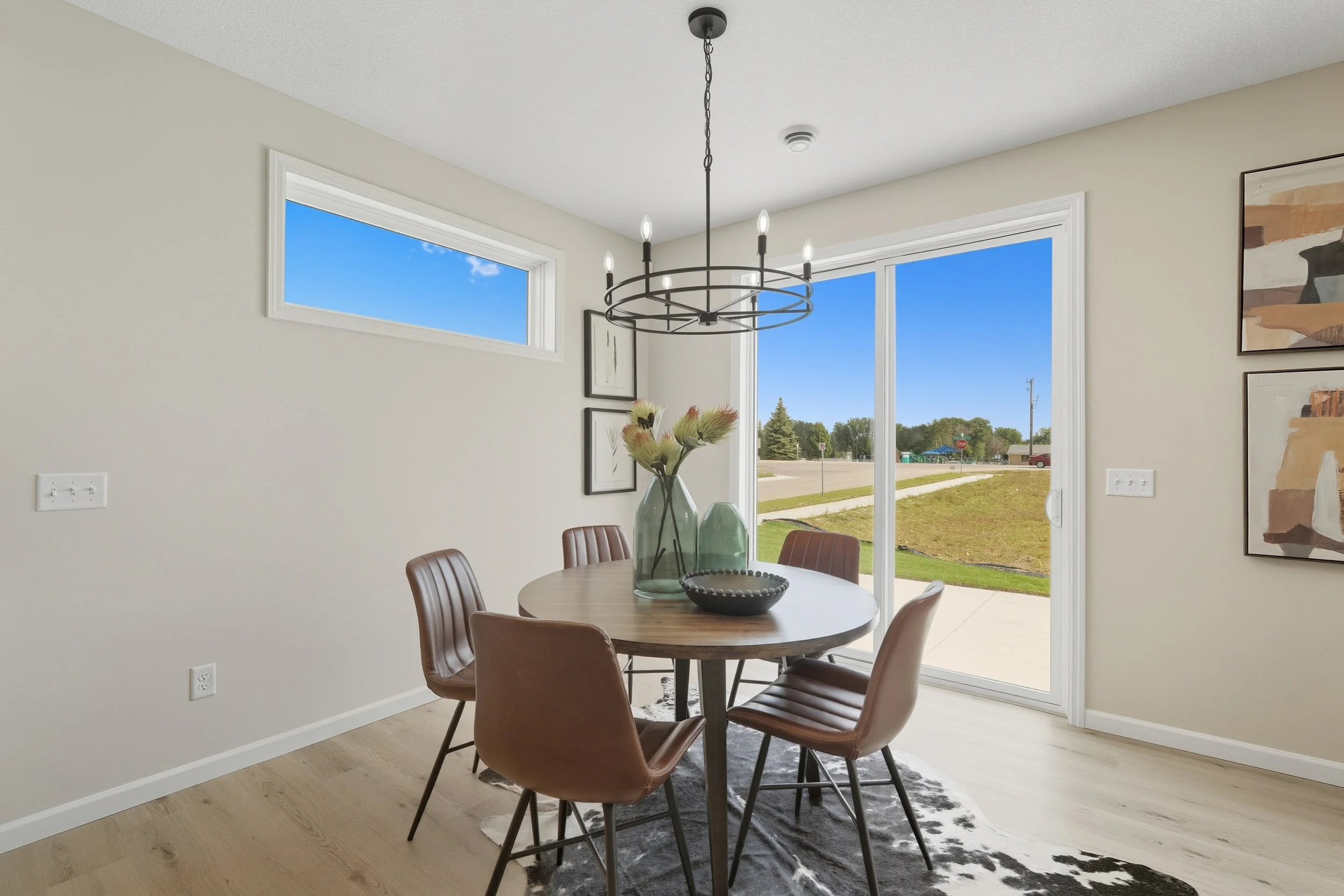 A modern dining room with a round wooden table, six brown chairs, and large windows showing a sunny outdoor scene. A black chandelier hangs above the table, which is decorated with vases and a bowl. The walls have minimalist artwork, and the room has