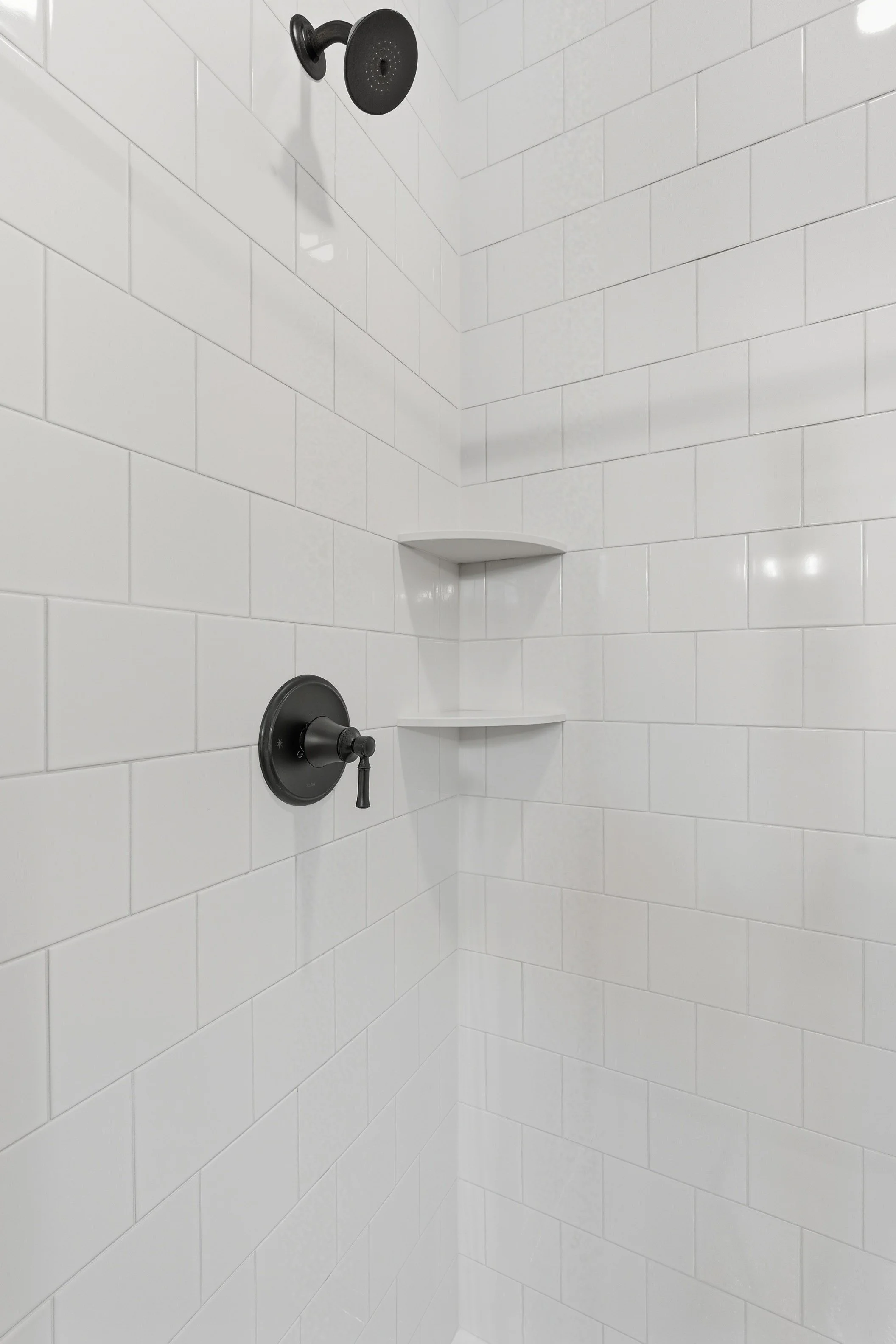 A white tiled shower with black showerhead and handle, featuring built-in corner shelves.