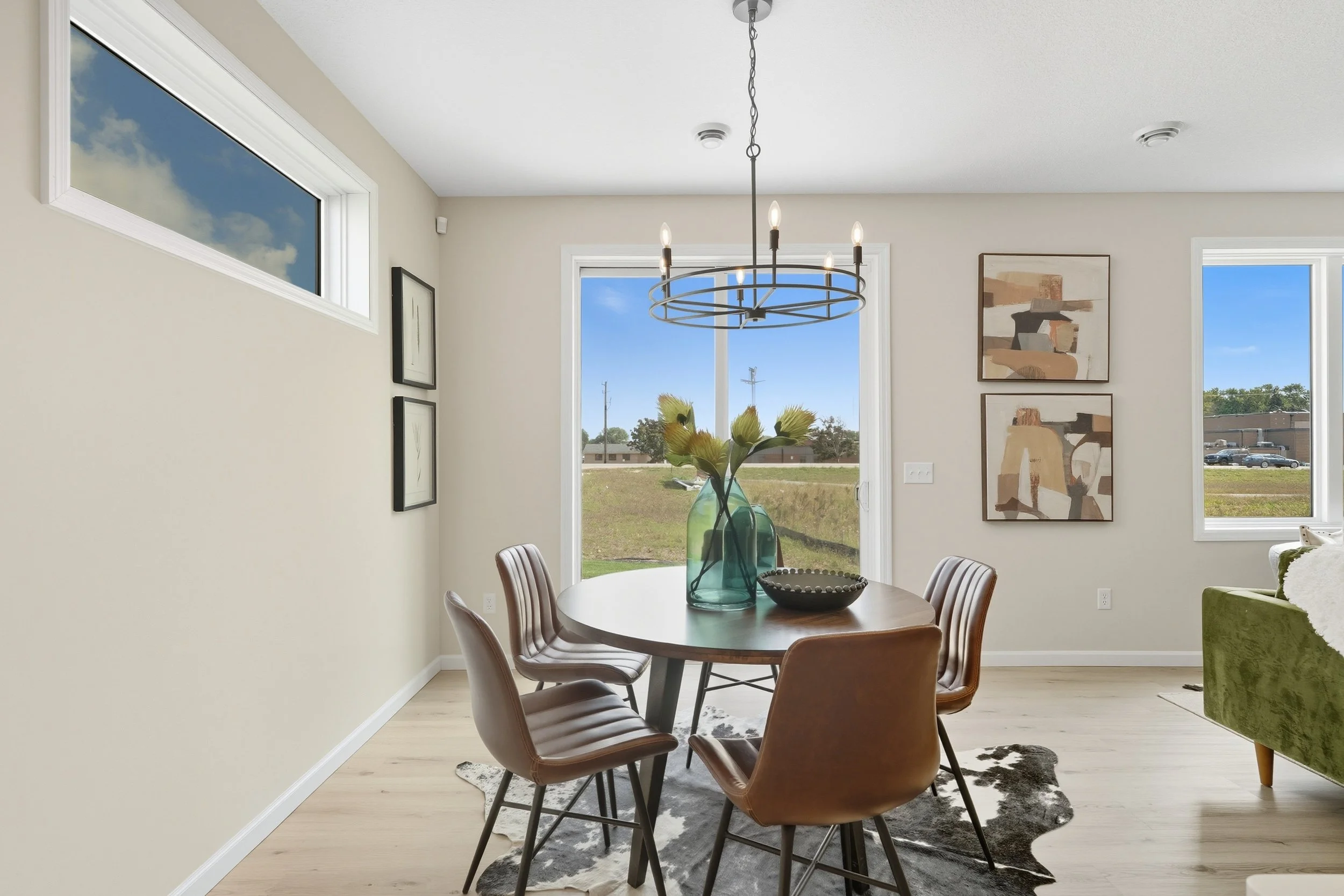 A dining room with a round wooden table, six chairs, a large vase with flowers, a black bowl, modern artwork on white walls, large windows with a view of the outdoors, and a modern chandelier hanging from the ceiling.
