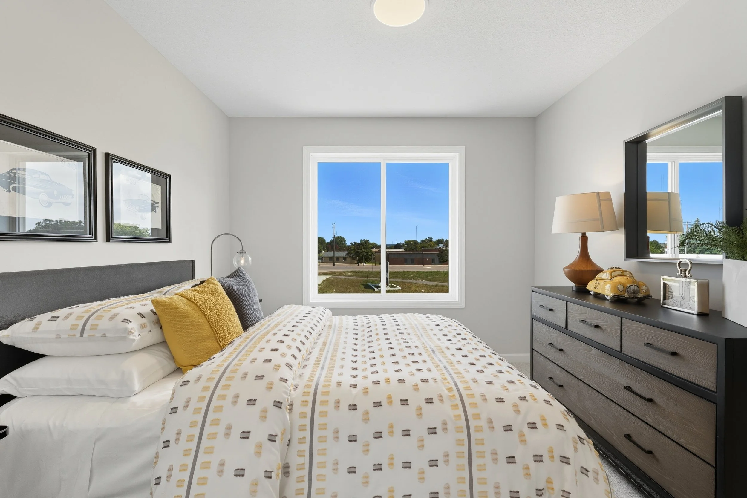 A tidy bedroom with a window showing a suburban neighborhood and a bright blue sky. The bed has white and beige patterned bedding with yellow and gray accent pillows. A dark dresser with a lamp, decorative yellow car, and photo frame is on the right.