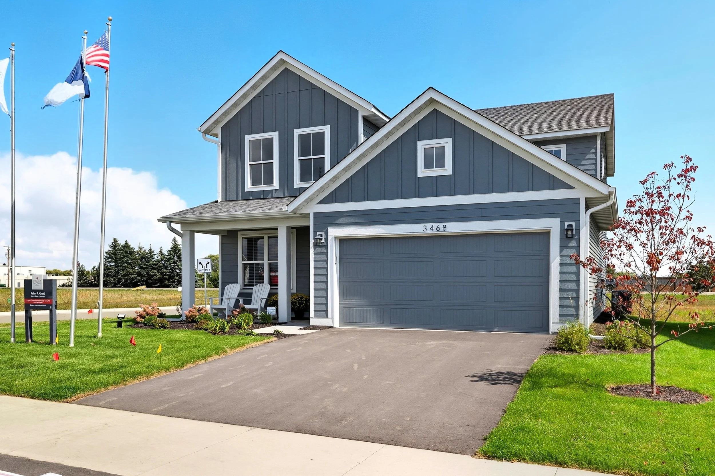 A two-story modern house with blue siding, white trim, a gray garage door, and a small front porch with chairs, surrounded by a green lawn and young trees, under a blue sky with scattered clouds.