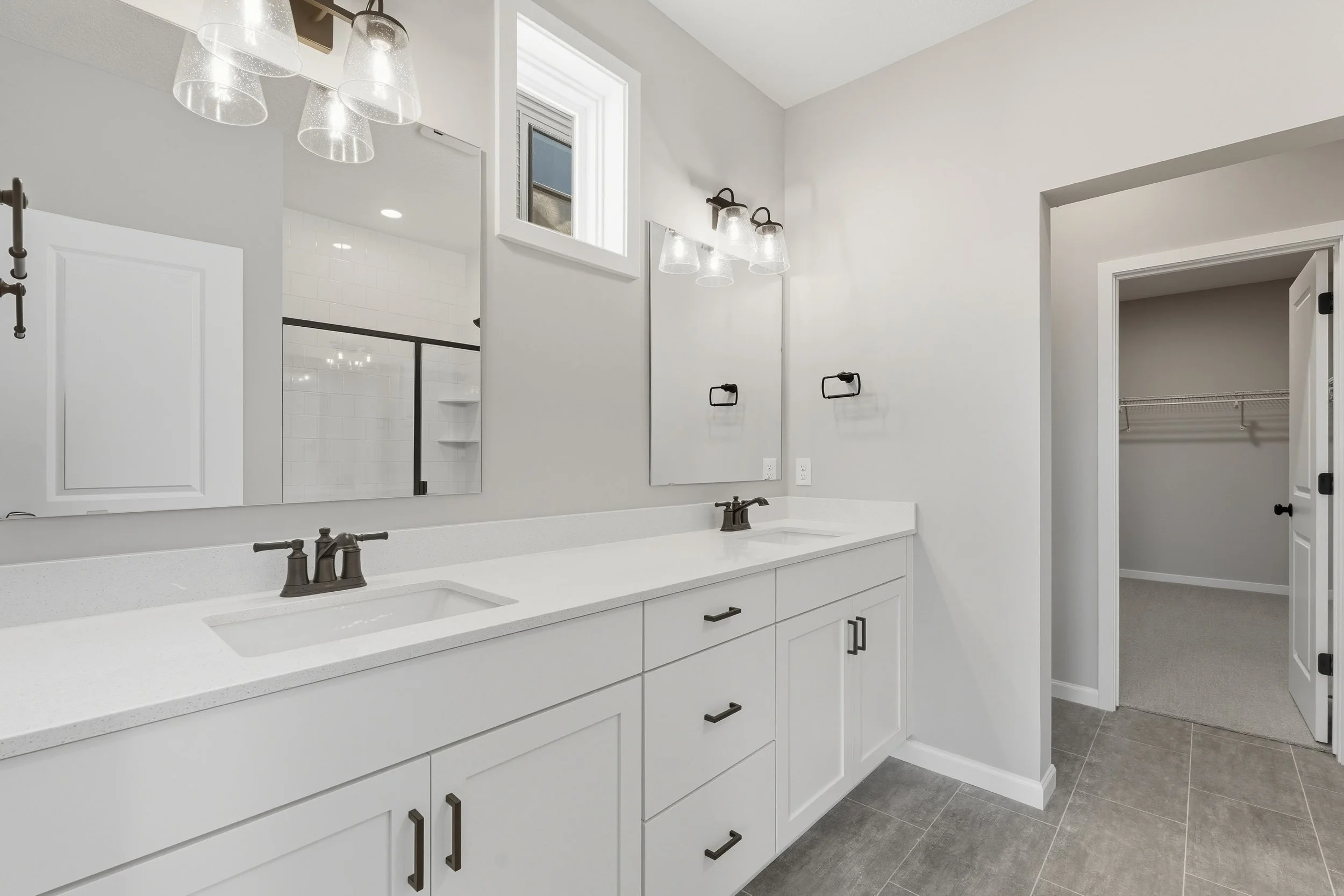 Modern bathroom with double vanity, white cabinets, black hardware, large mirror, wall-mounted light fixtures, small window, and an open closet with wire shelving.
