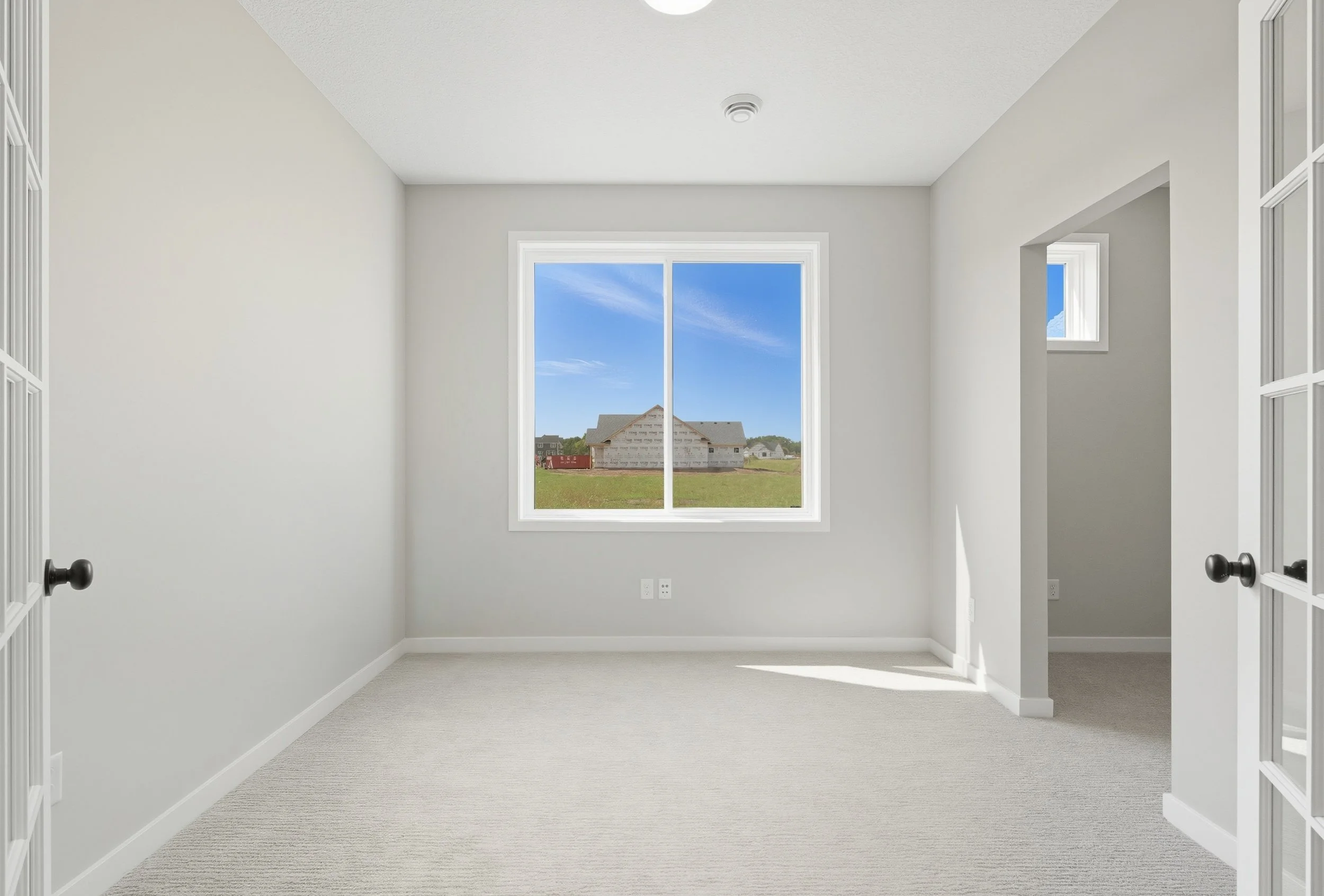 Empty room with white walls, beige carpet, large window showing a house under construction outside, and smaller window in an adjacent wall.