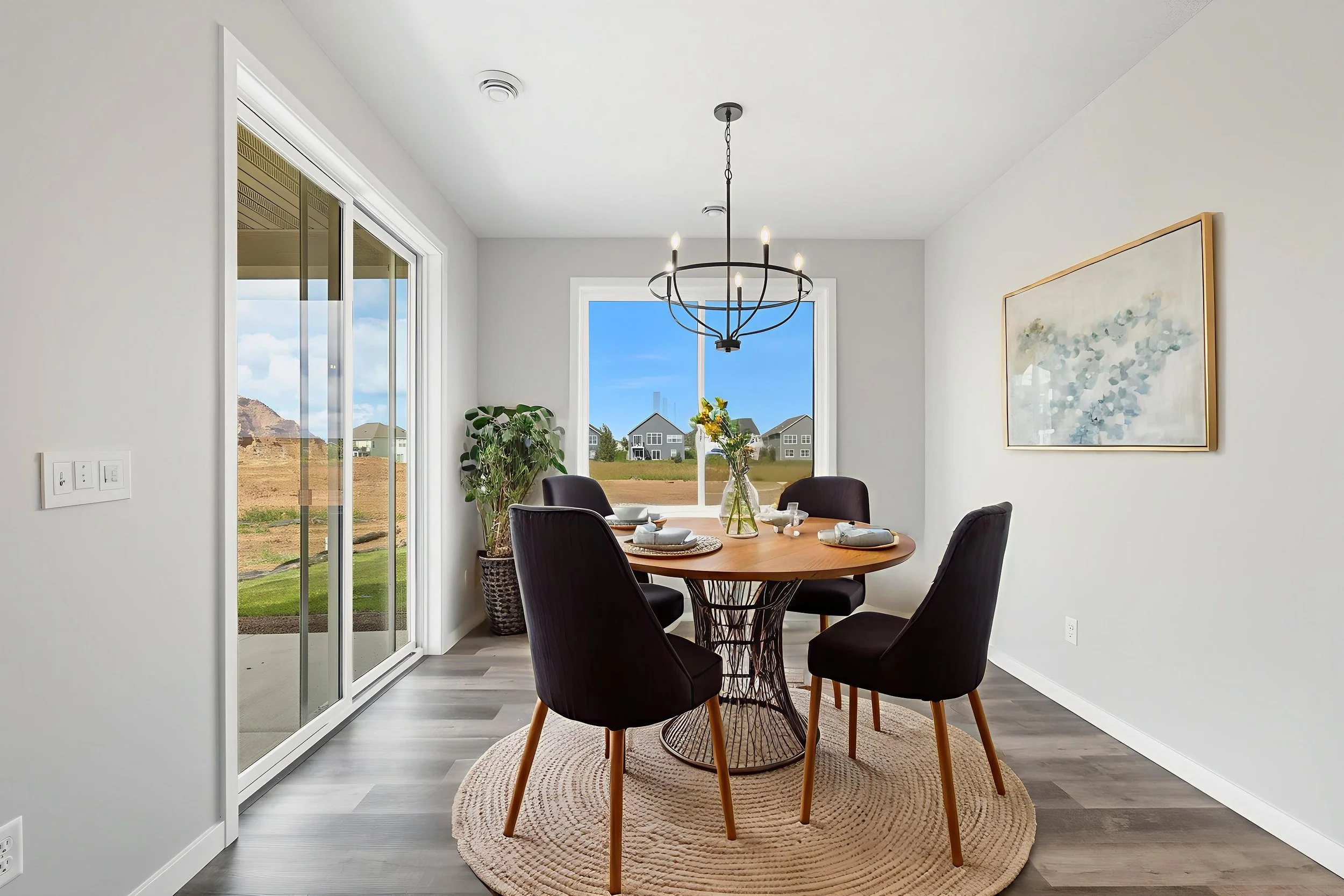A modern dining room with a round wooden table, four black chairs, a light fixture hanging above, a large window with a view of a neighborhood and blue sky, a sliding glass door, a potted plant, a framed abstract painting on the wall, and a round rug