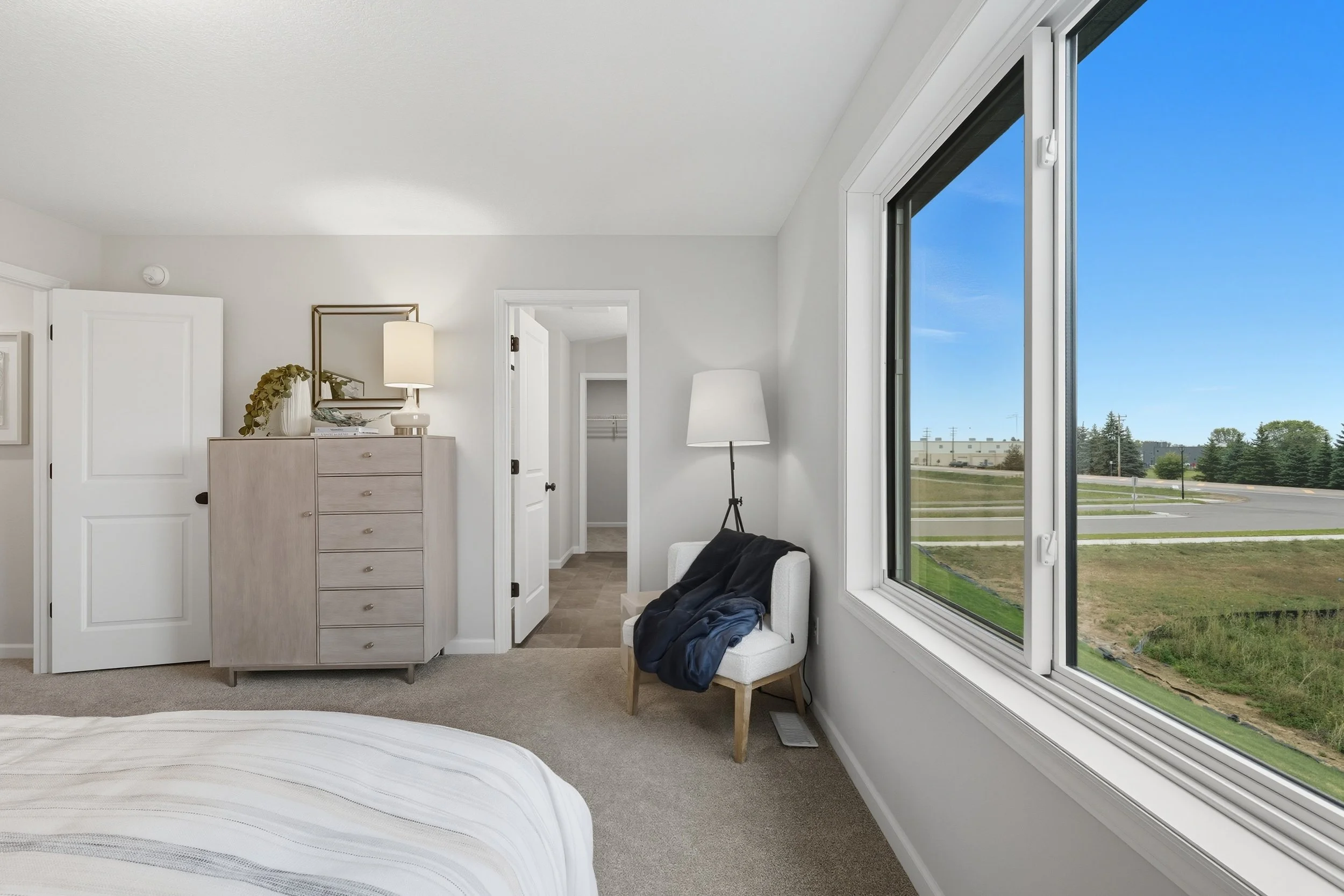 A bedroom with a large window showing a view of green grass and a parking lot, a white armchair with a black jacket on it, a lamp next to the chair, a gray dresser with a mirror and a lamp, and an open closet door.