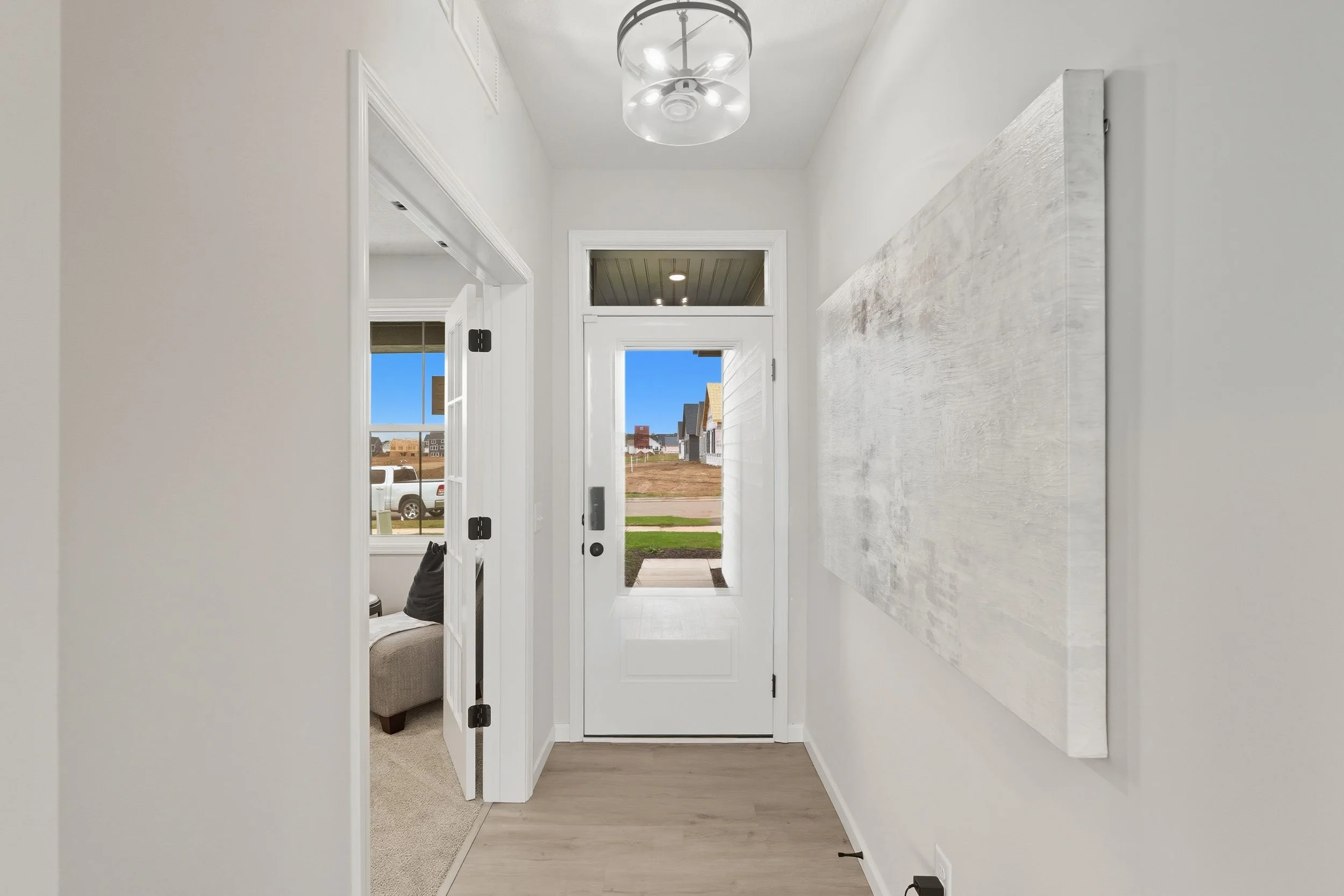 Bright entryway with a white front door with glass panel, artwork on the right wall, and a bedroom visible through the doorway on the left.
