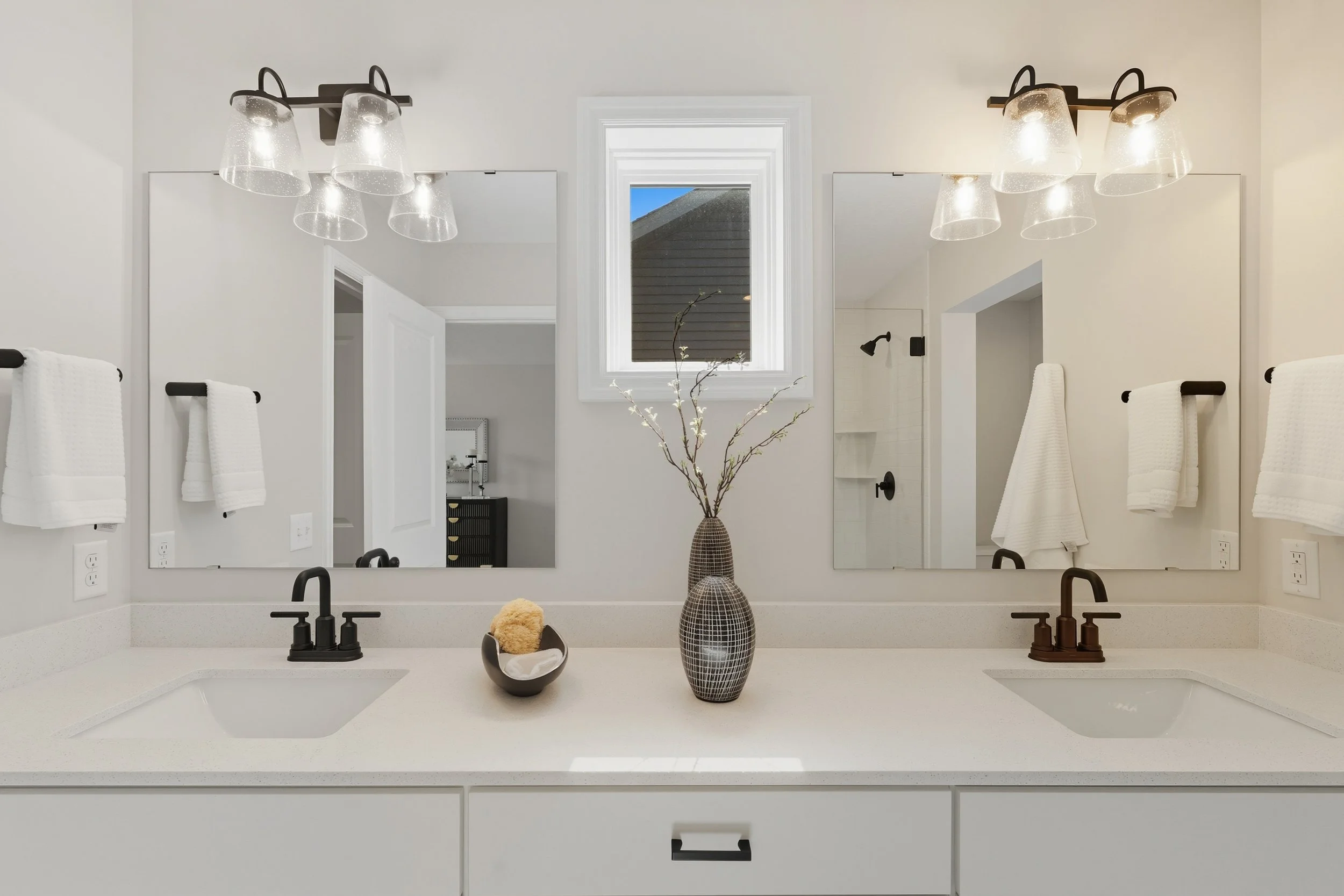 Modern bathroom with double sink vanity, large mirror, wall-mounted light fixtures, white towels, decorative vases, and a window above the sinks.