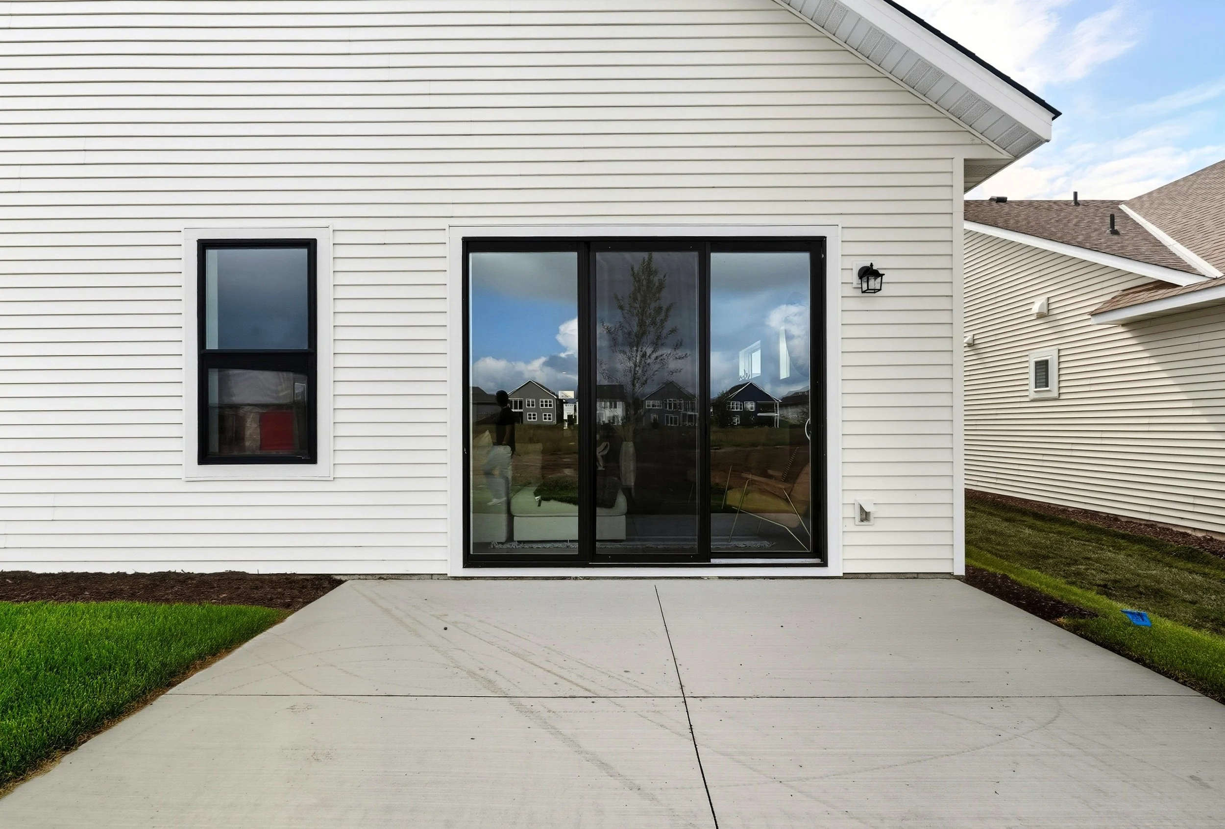 View of the back of a house with white vinyl siding, a large sliding glass door, a small window, an outdoor light fixture, and a concrete patio surrounded by grass and neighboring houses in the distance under a partly cloudy sky.