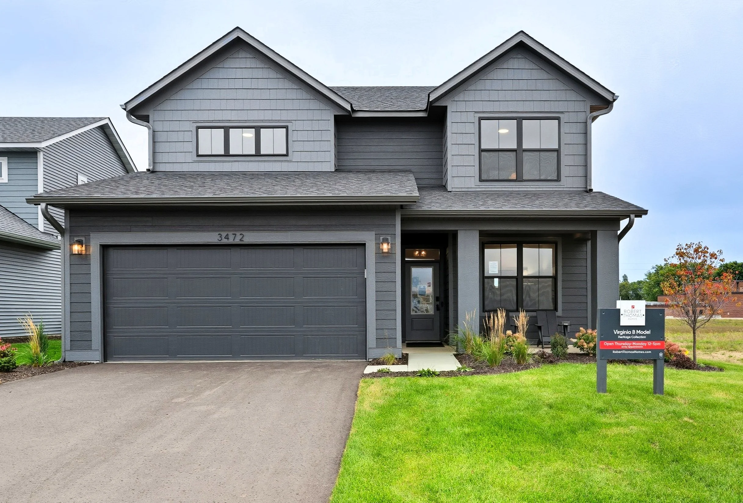 A modern two-story house painted in dark gray with black window frames and a matching garage door. The front yard has a well-maintained lawn, small plants, and a sidewalk leading to the front door. A sign in the yard indicates the house is for sale b