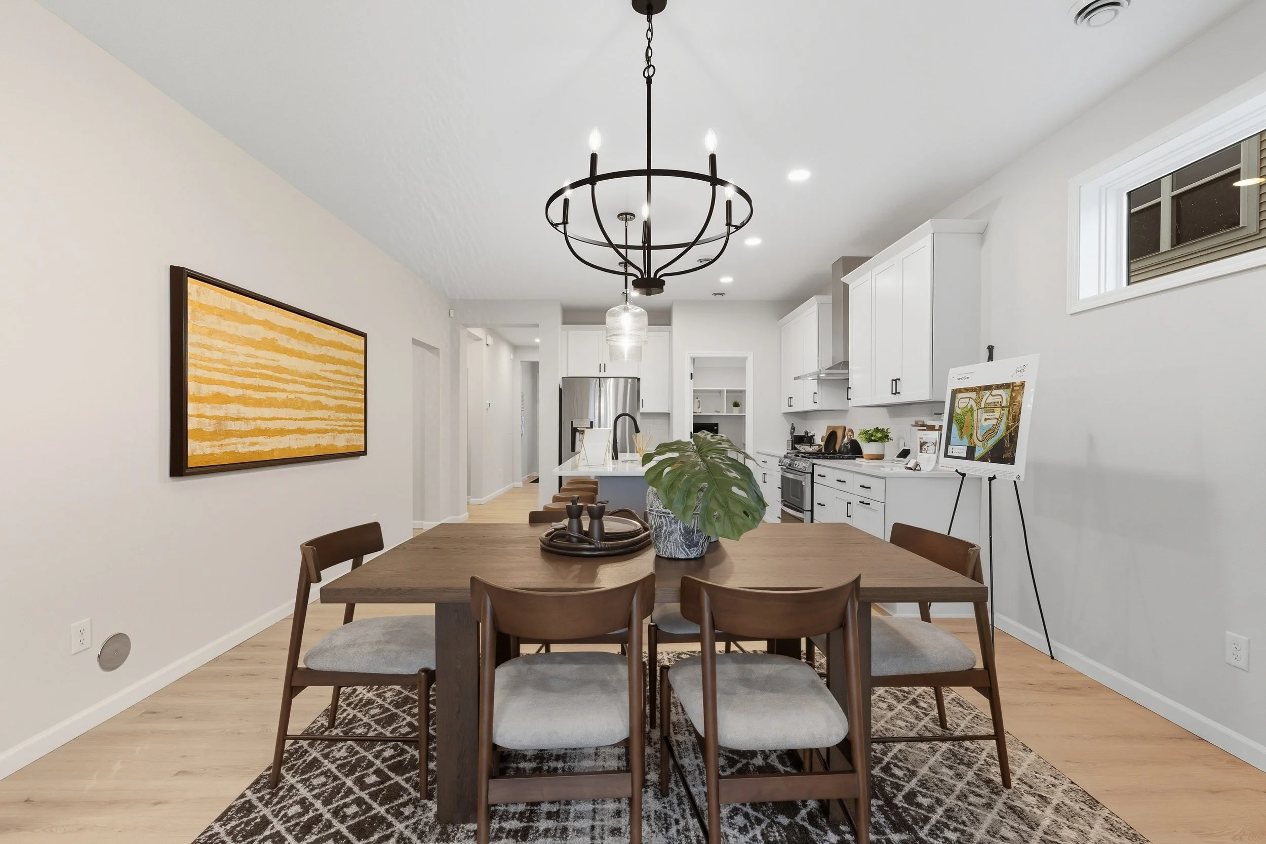 Dining room with a wooden table, six chairs, a large green leafy plant as centerpiece, modern chandelier, white kitchen with stainless steel appliances, artwork on wall, and a small high window.
