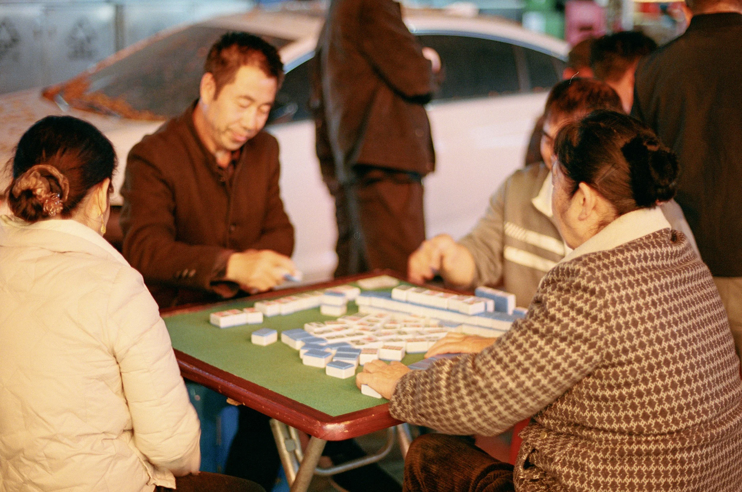 A group of elderly people playing Mahjong outdoors at night, sitting around a green table with Mahjong tiles.