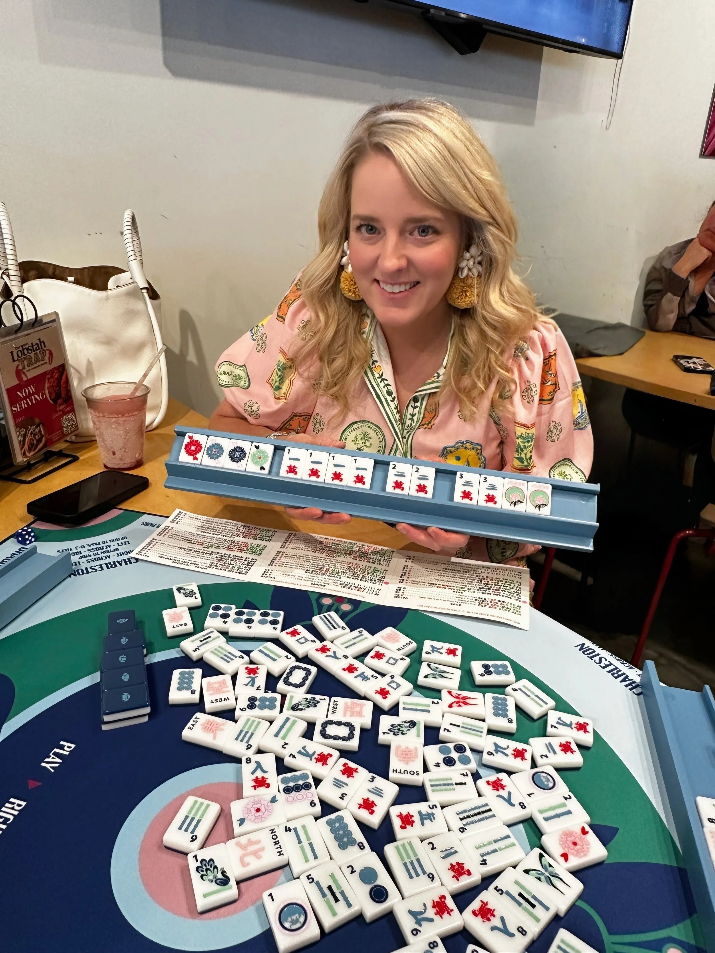 A woman with blonde hair, earrings, and a pink patterned shirt sitting at a table playing Mahjong, with Mahjong tiles on the table and a rack of tiles in her hands, in a casual restaurant setting.