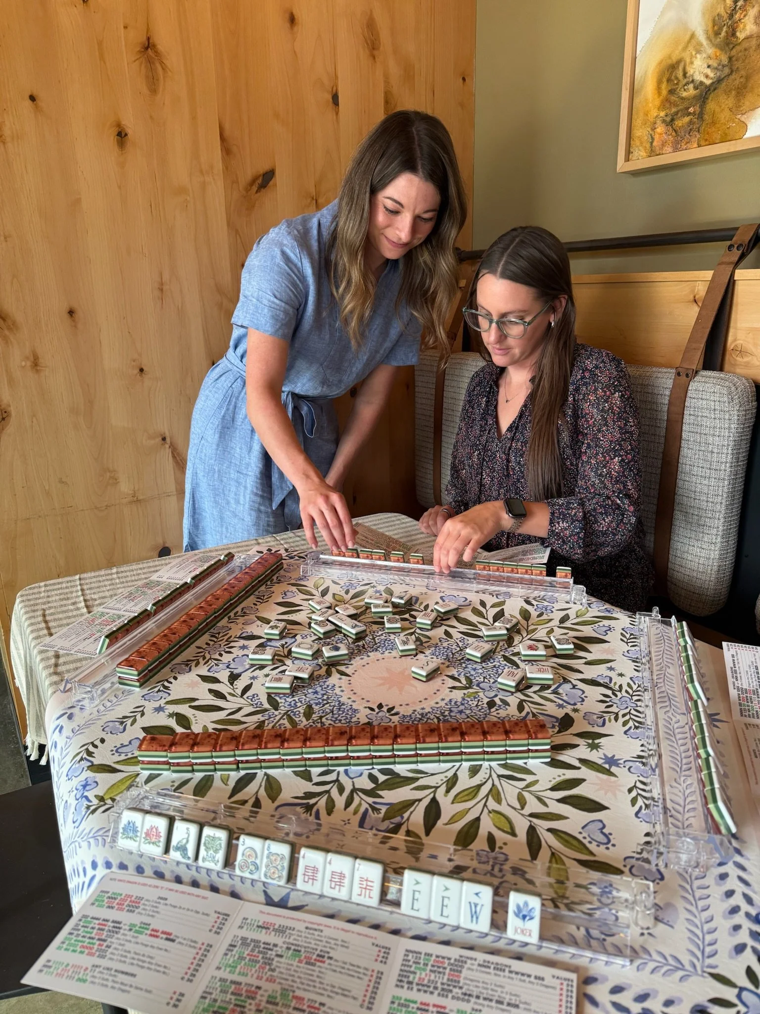 Two women playing Mahjong at a table with game tiles and score sheets.