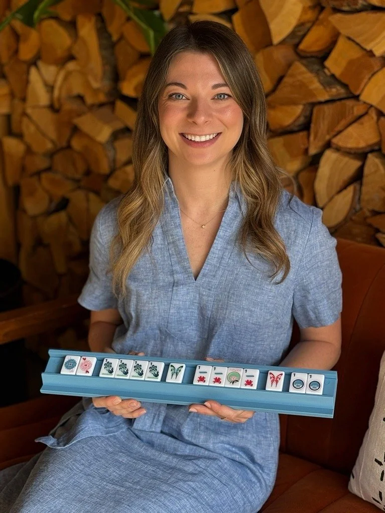 A woman with long hair wearing a light blue dress, smiling and sitting on a couch, holding a rack with multiple mahjong tiles. The background features a wood log wall.