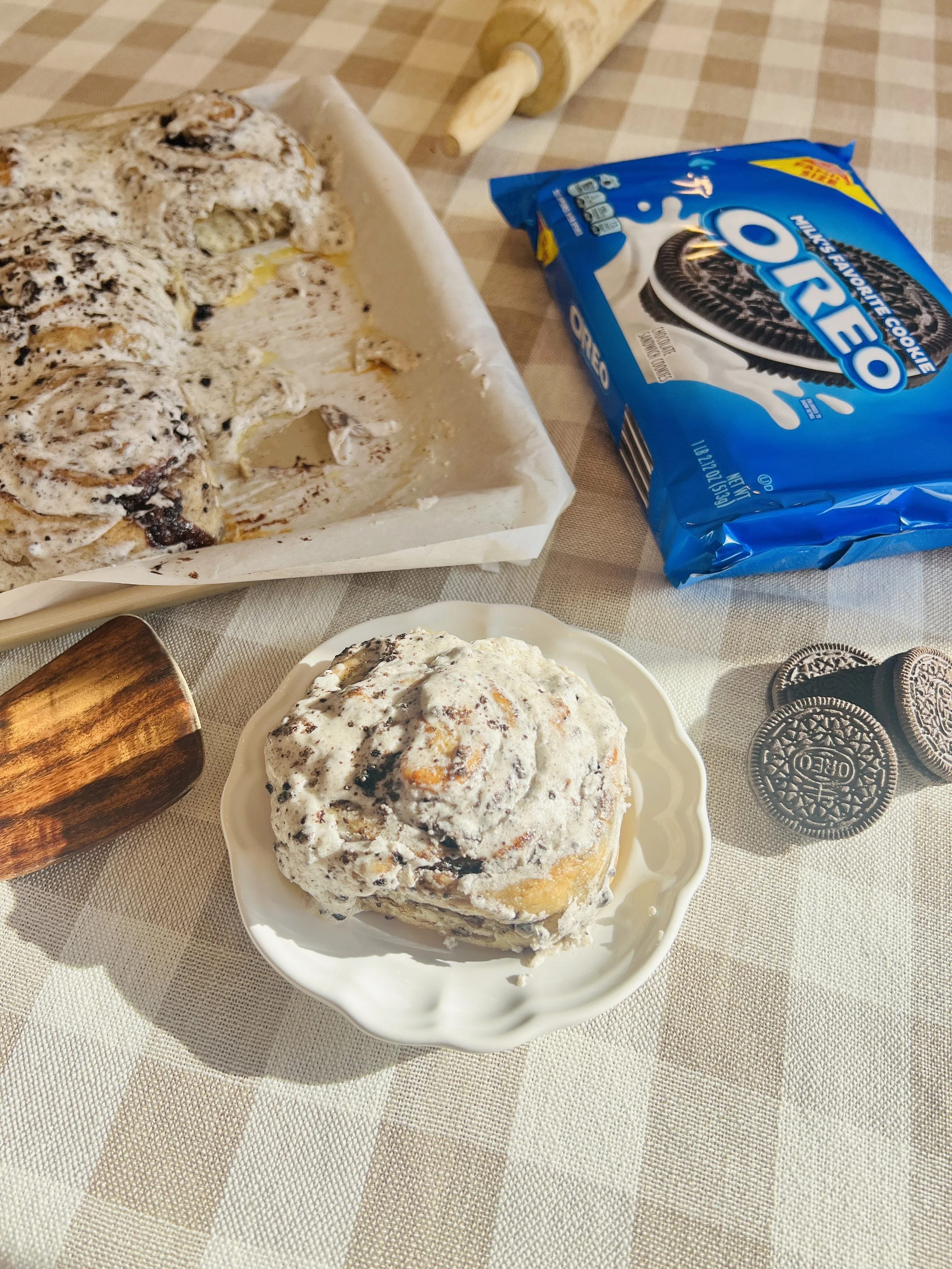 A plate with a scoop of cookies and cream ice cream, a baking tray with partially eaten cookies, a bag of Oreo cookies, a wooden rolling pin, and a few whole Oreo cookies on a checkered tablecloth.