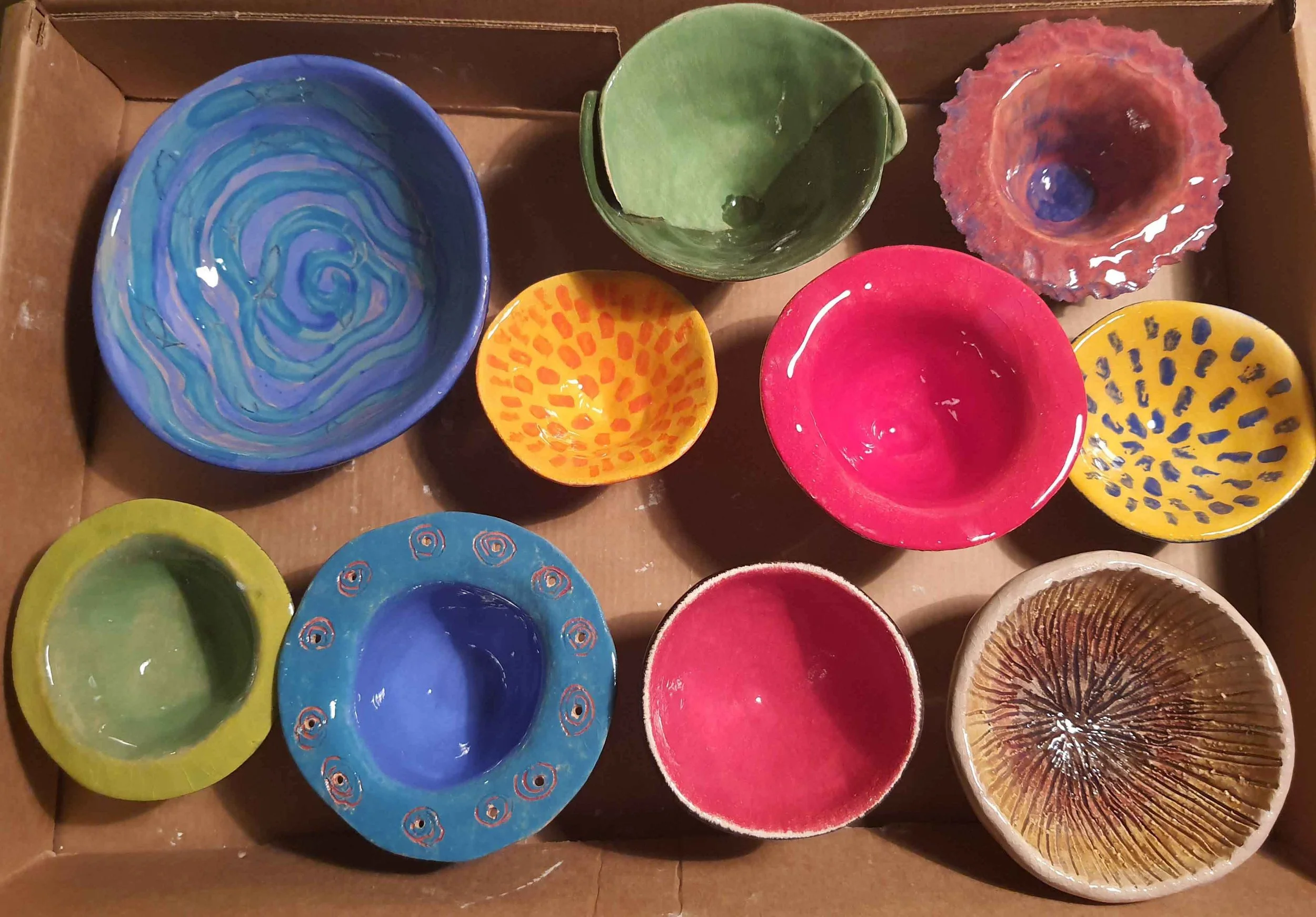 Colored ceramic bowls and cups in a cardboard box, with patterns and textures including swirls, spots, and ruffles.