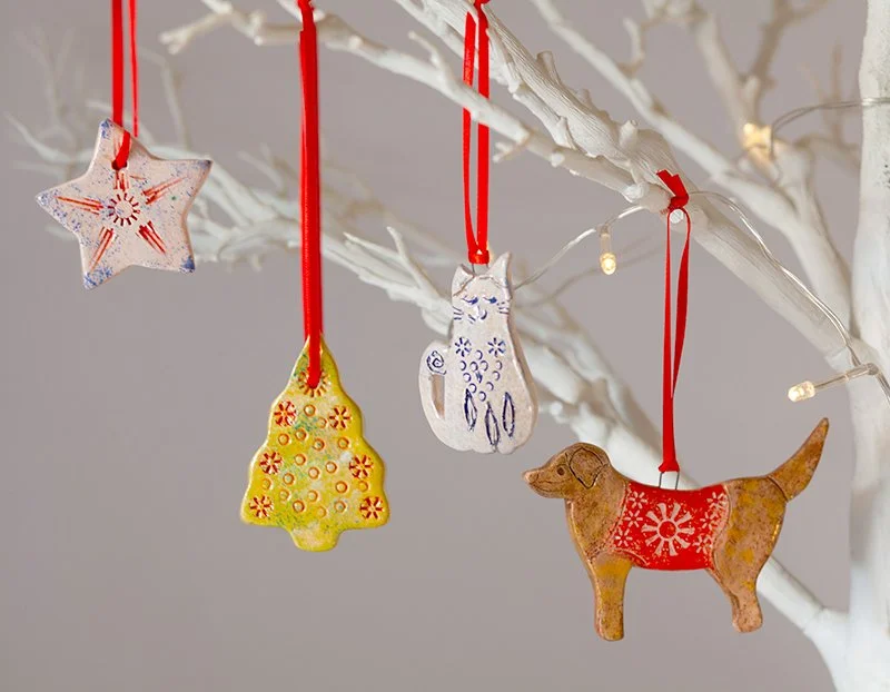 Four holiday ornaments hanging from a white decorated tree branch with red ribbons, including a star, a tree, a cat, and a dog.