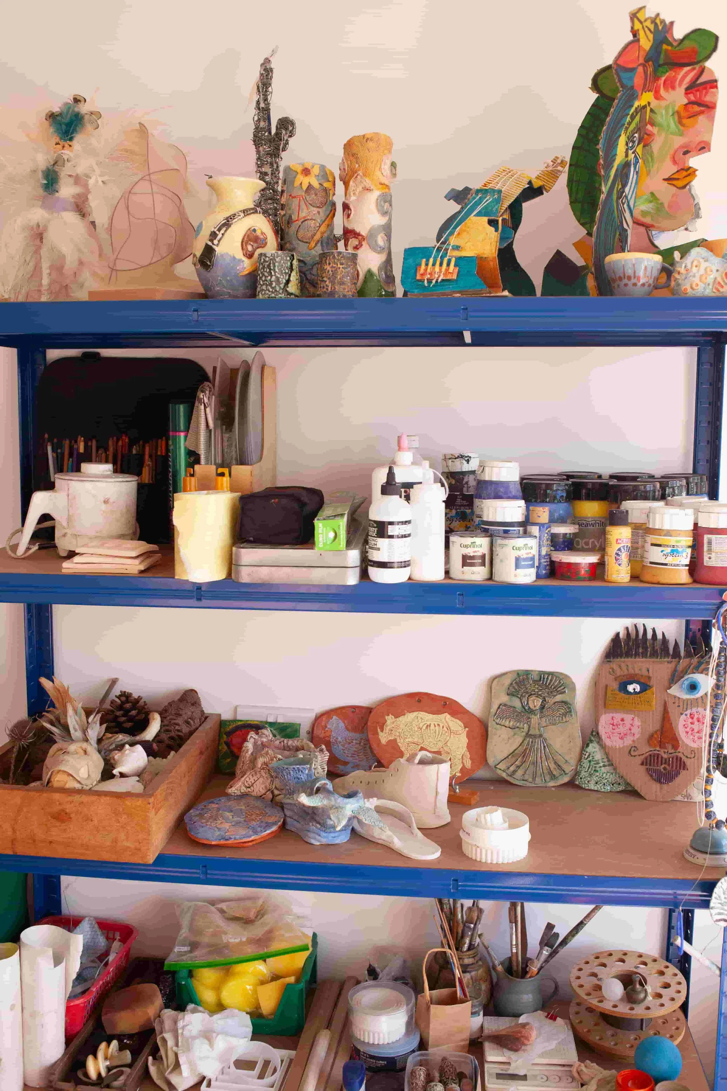 A blue metal shelving unit filled with art supplies, ceramics, and decorative items, including painted ceramics and handmade crafts.