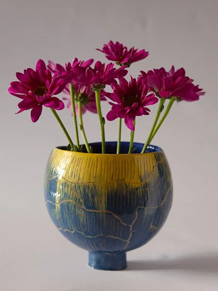 A ceramic vase with yellow and blue glaze holding pink and purple flowers.