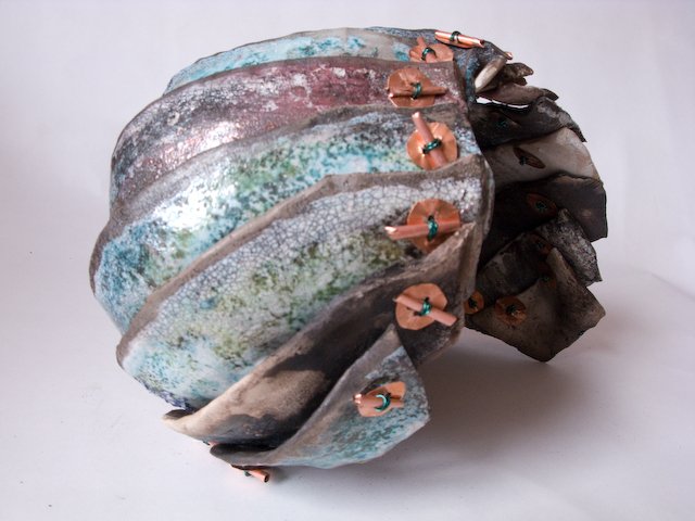 An abstract mixed media sculpture resembling a large, textured oval shape with metallic and painted surfaces, adorned with small copper-colored sculptures attached with green elements.