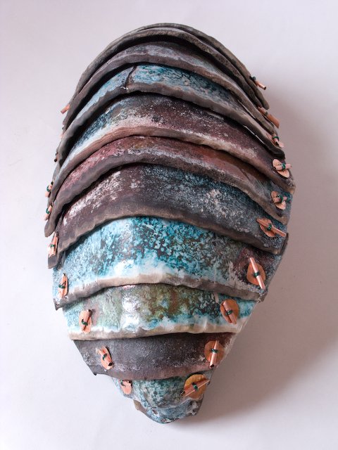 Close-up of a layered, weathered, earth-toned acorn shell with small copper-colored pins attached to each layer.
