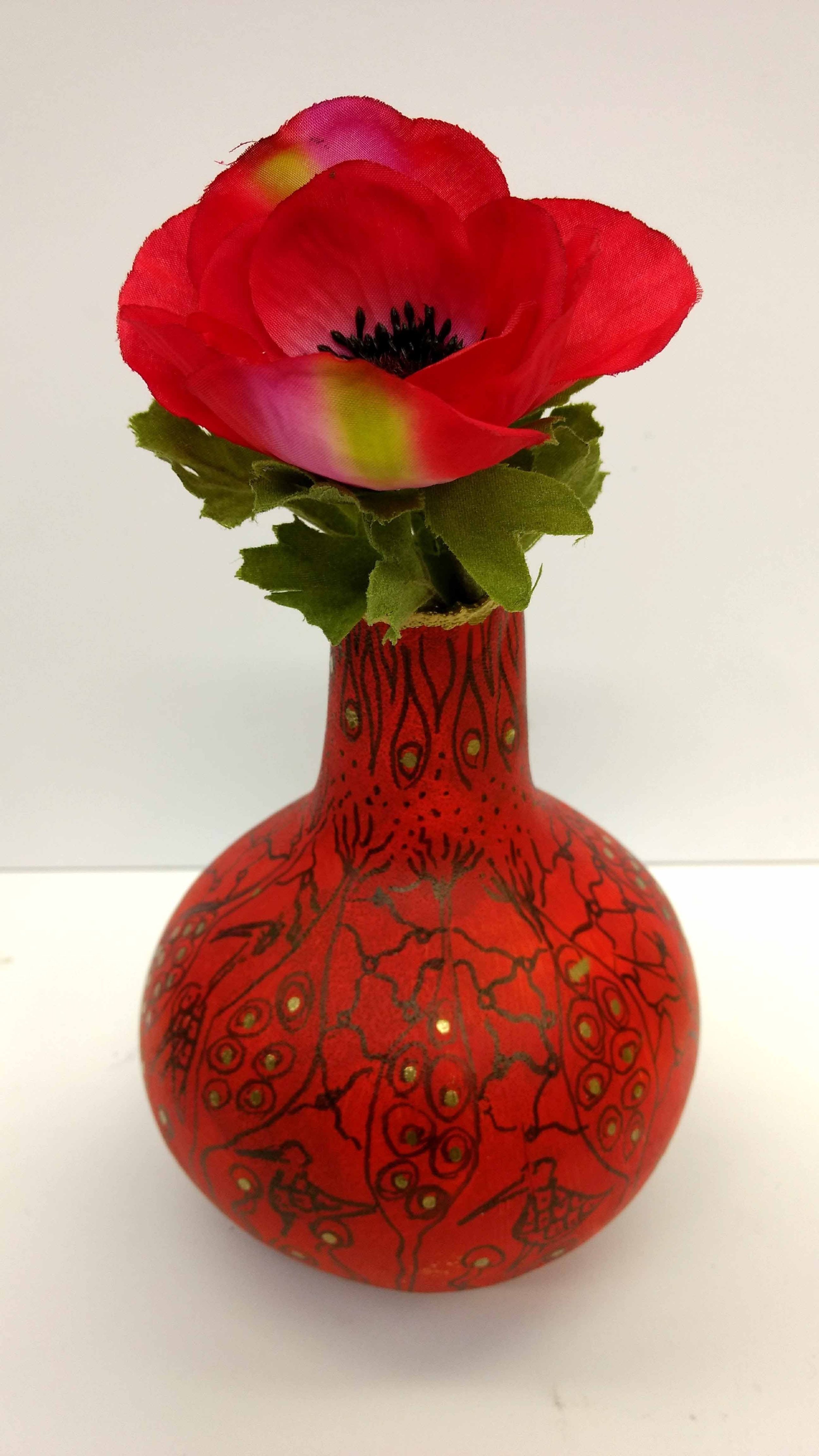 A red decorative vase with black and gold patterns holding a vibrant red artificial flower with green leaves.
