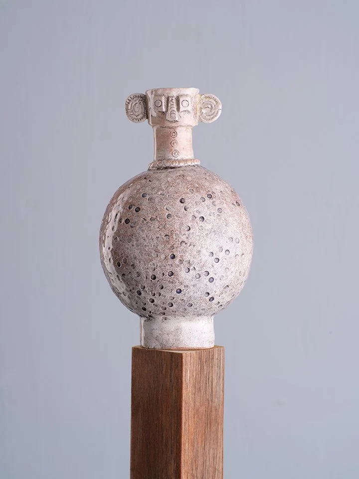 A ceramic sculpture of a humanoid head with prominent facial features and large ears, mounted on a spherical body with small circular perforations, supported by a wooden pedestal.