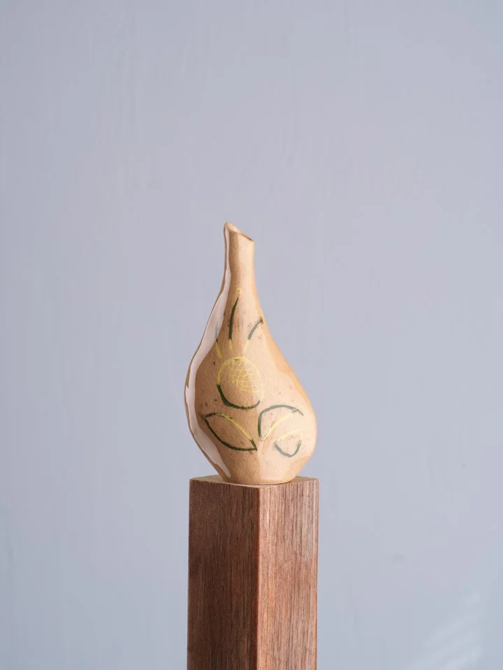 Ceramic vase with a beige glaze and painted abstract green and yellow designs, placed on a wooden pedestal against a plain gray background.