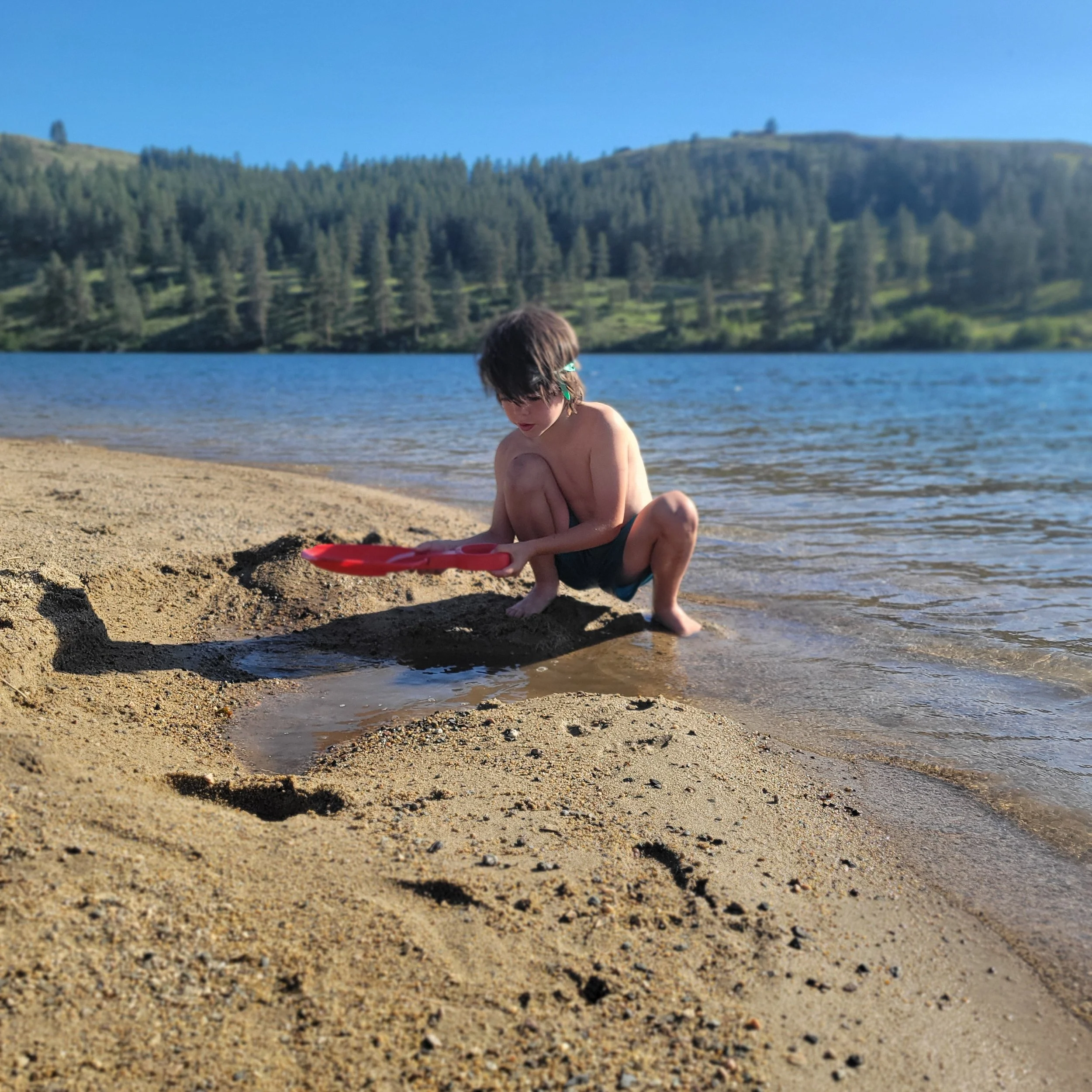 A young boy playing with a red shovel in the sand at a lakeside beach with mountains and trees in the background.