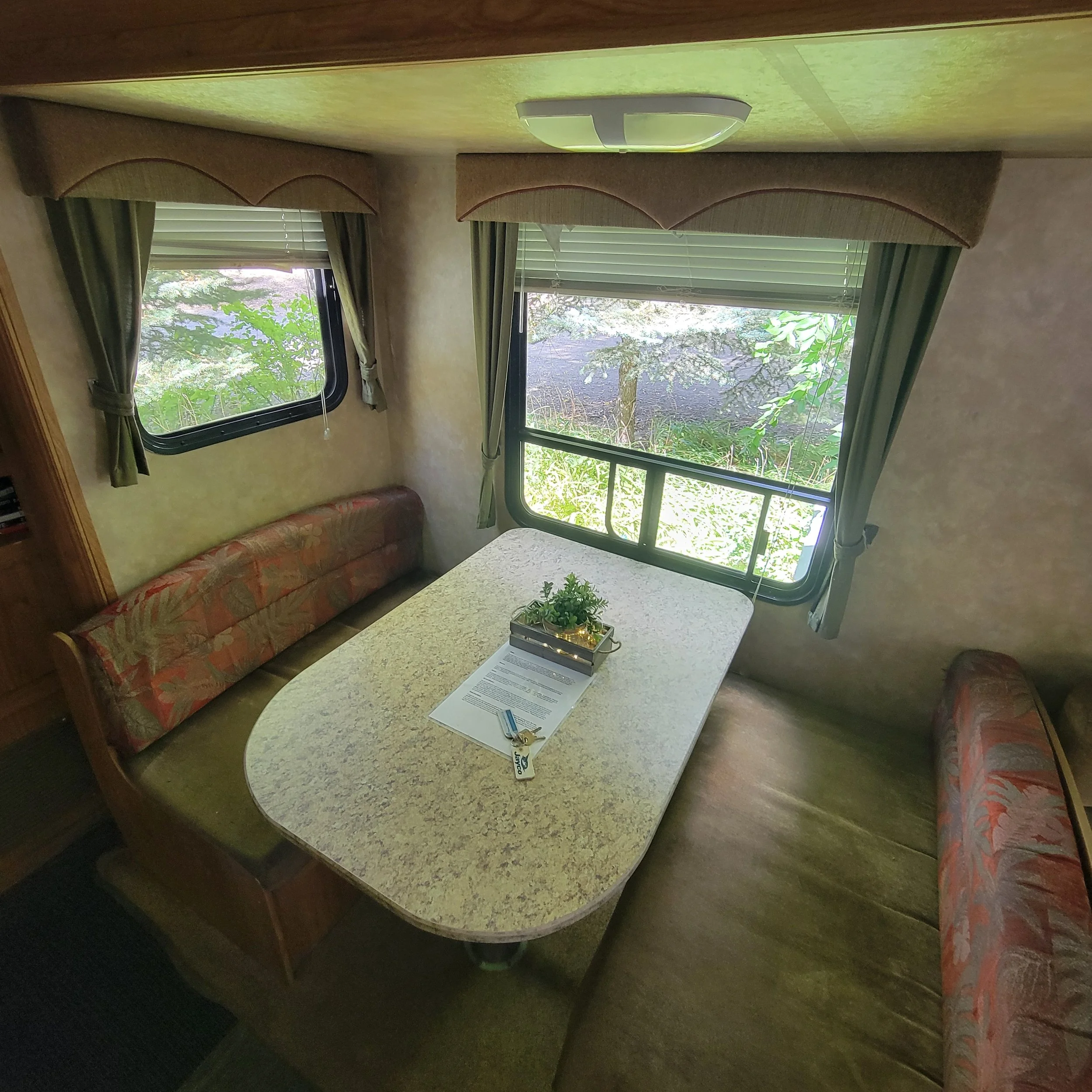 Interior view of a dining area with a table, two benches, two windows with curtains, and a ceiling light fixture.
