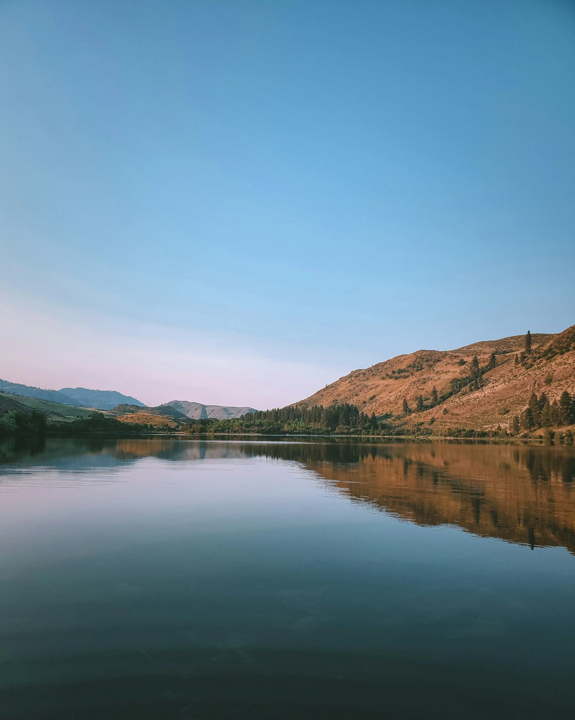 Calm lake reflecting surrounding hills and trees under a clear blue sky.