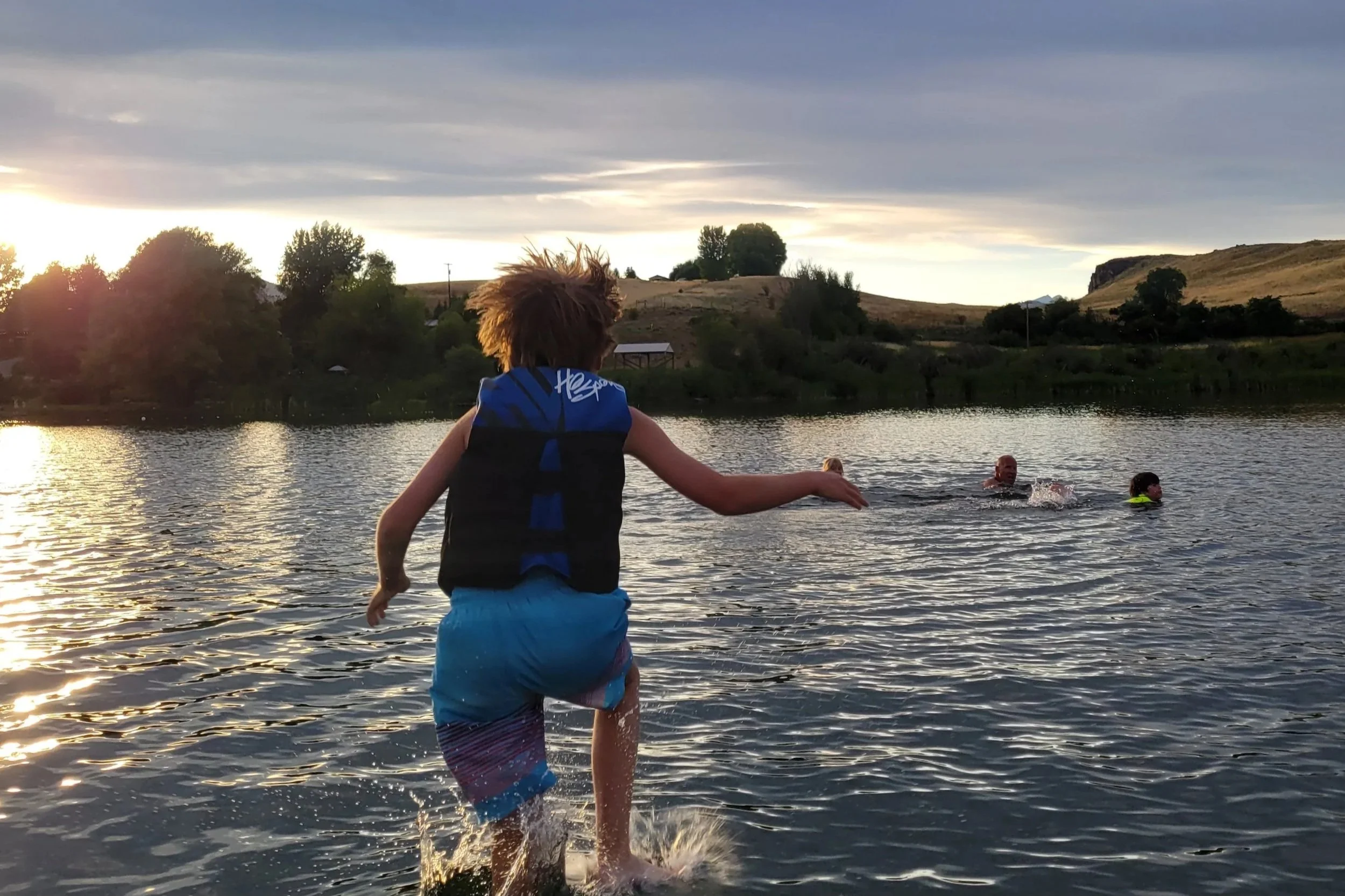 Children playing and swimming in a lake during sunset, with hills and trees in the background.