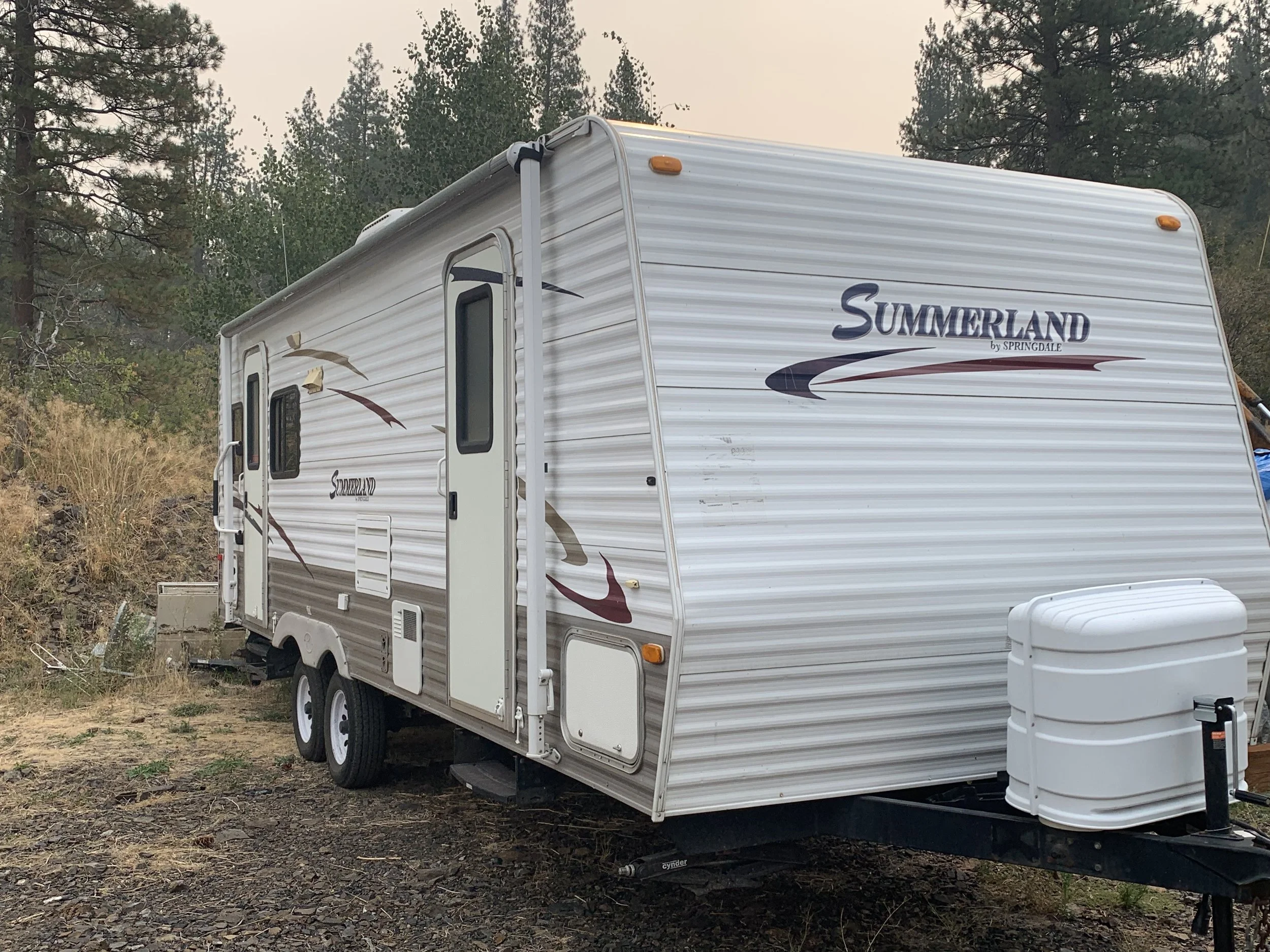A white travel trailer named 'Summerland' parked on dirt ground with trees and bushes in the background.
