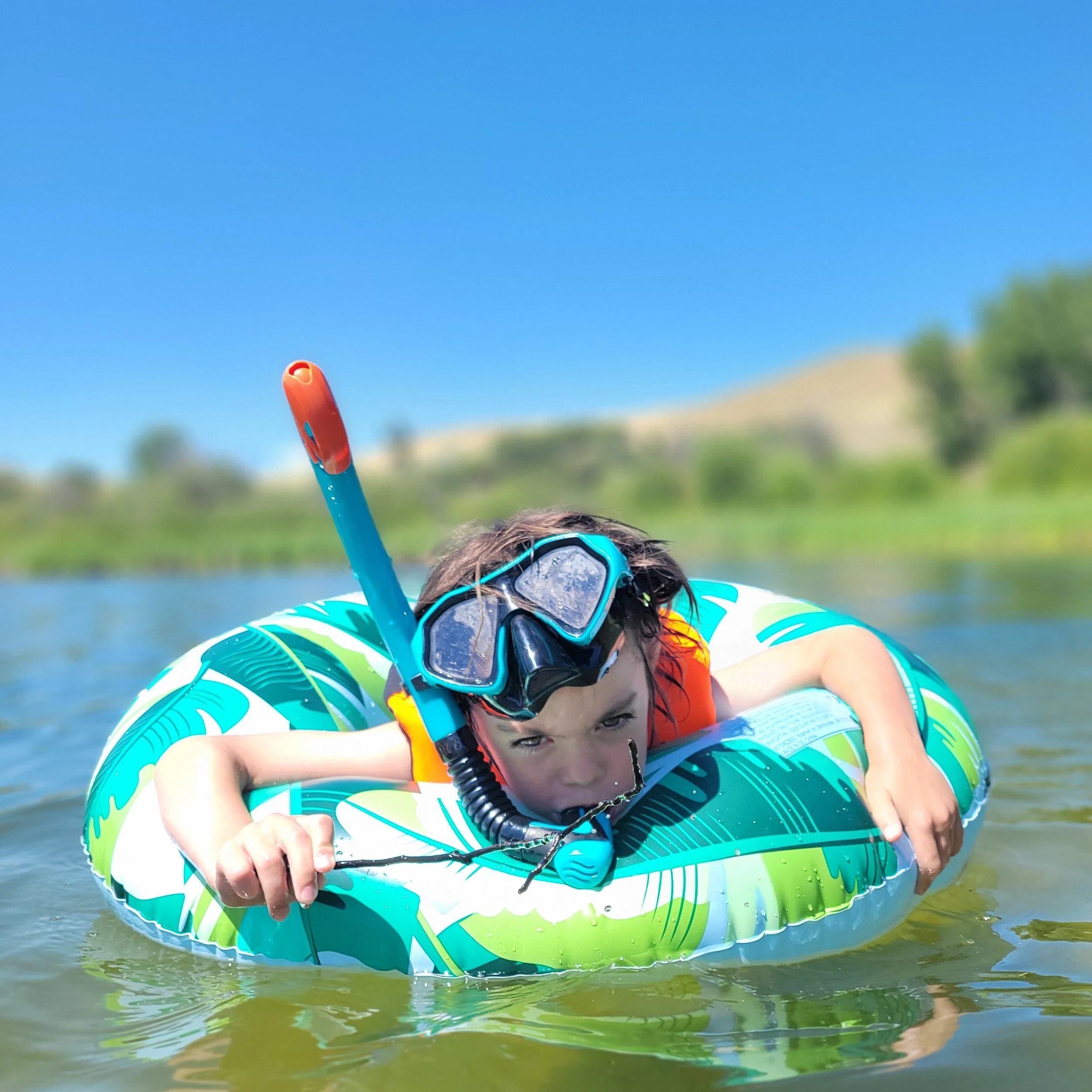 Child floating on a colorful inflatable ring in a lake, wearing snorkeling gear and holding a snorkel, with green hills and blue sky in the background.