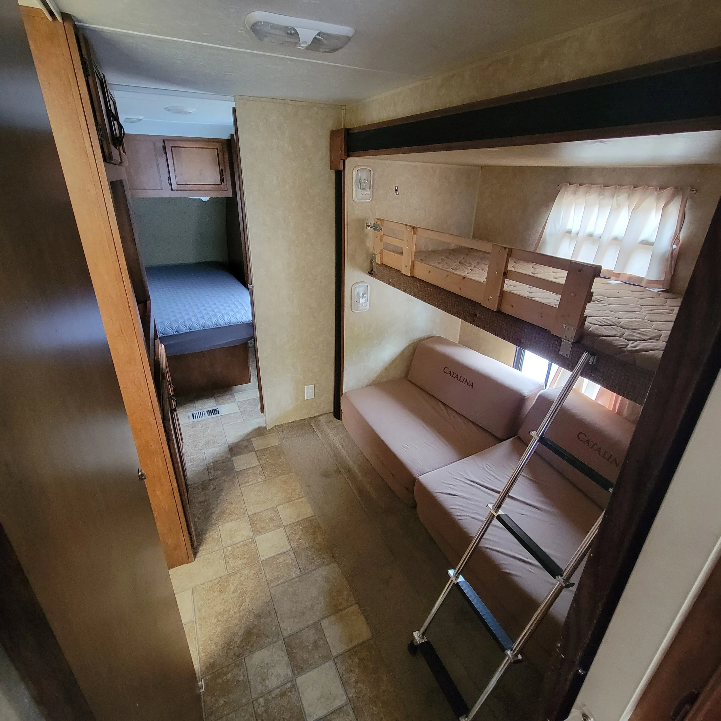 Interior of a small living space with a bunk bed above a beige fold-out sofa, a ladder leaning against the sofa, a window with curtains, and a doorway leading to a small bedroom with a single bed and wooden cabinets.