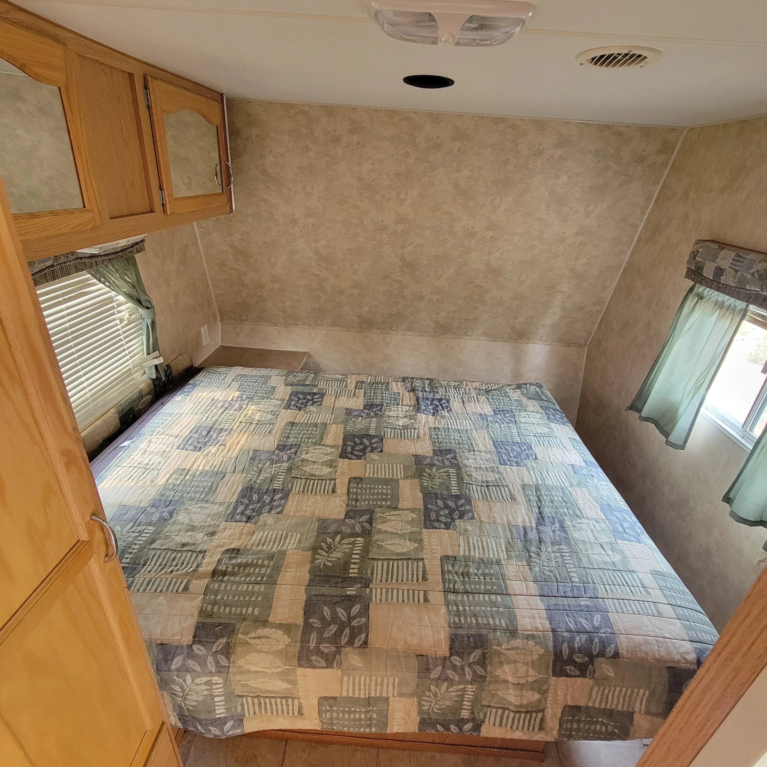 A bedroom with a double bed with a patchwork quilt, two windows with green curtains, and wooden cabinets in an RV or mobile home.