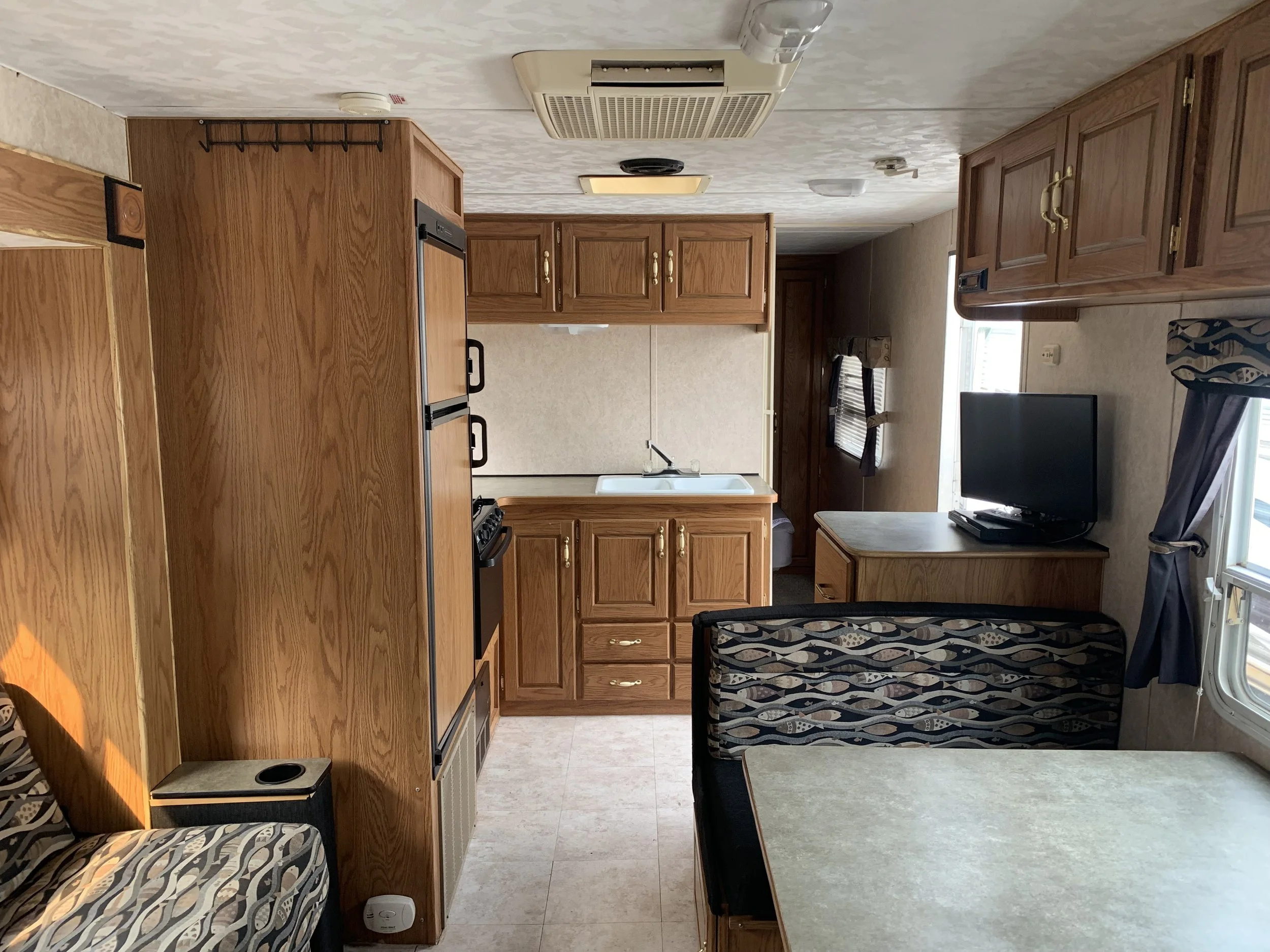 Interior of a small kitchen and dining area in a mobile home or RV with wood cabinets, a stove, a sink, a small television, and a table with a patterned curtain.