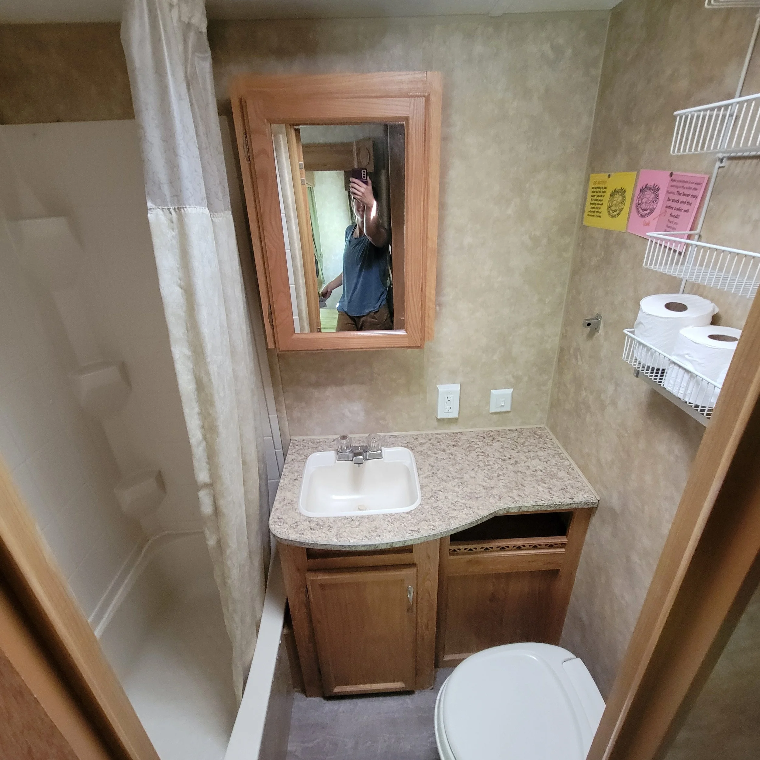 Small bathroom with a shower, a sink with a granite countertop, a mirror, a toilet, a wall-mounted wire shelf with toilet paper rolls, colorful signs, and a person taking a photo of the mirror reflection.