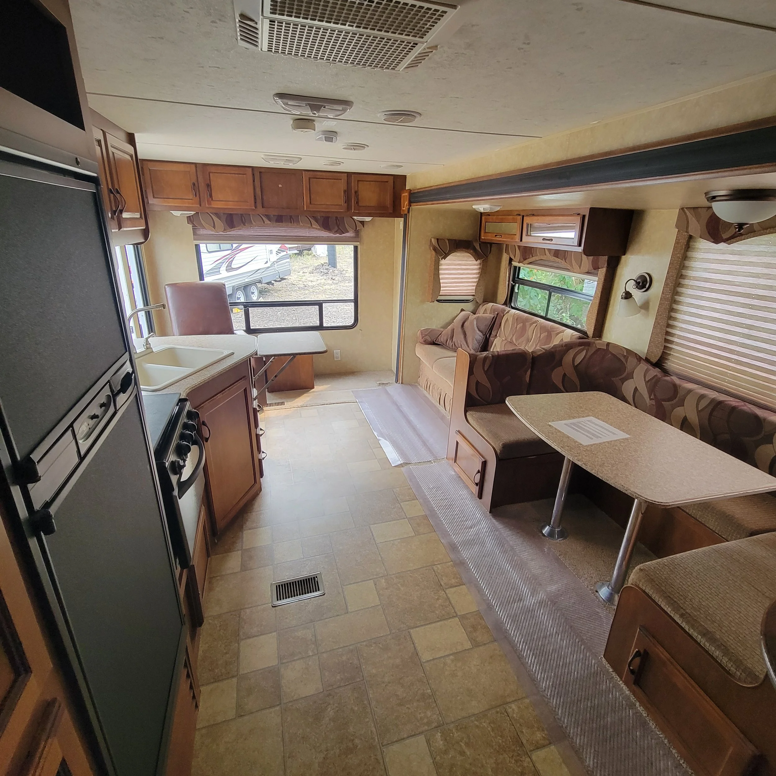 Interior of a recreational vehicle with a kitchen area on the left, a dining area on the right, and a living space with a sofa and windows in the background.