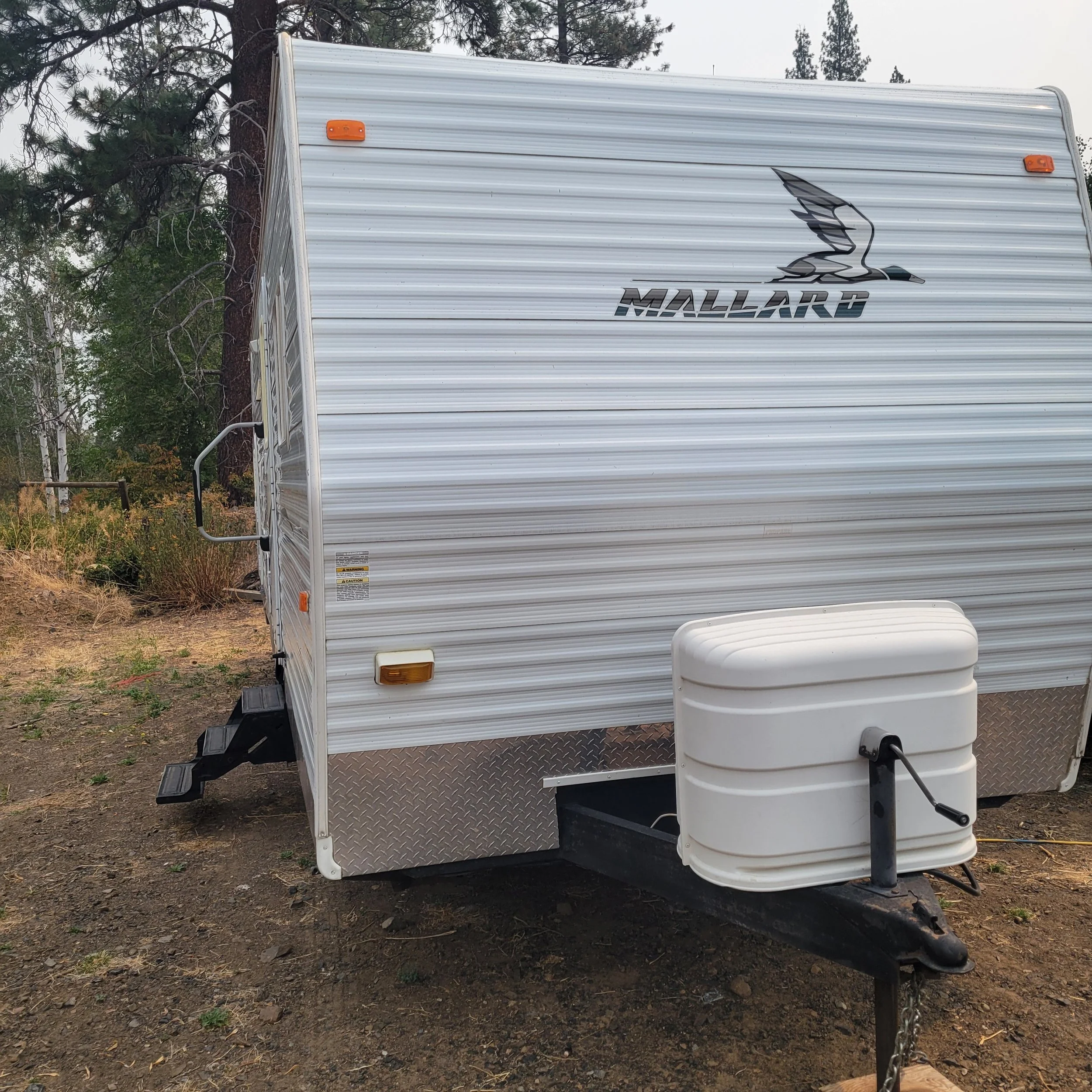 A white Mallard travel trailer with metal accents parked on dirt, featuring a logo of a flying mallard bird on the front. The trailer has small orange marker lights, a fold-out step, and a white propane tank at the front.