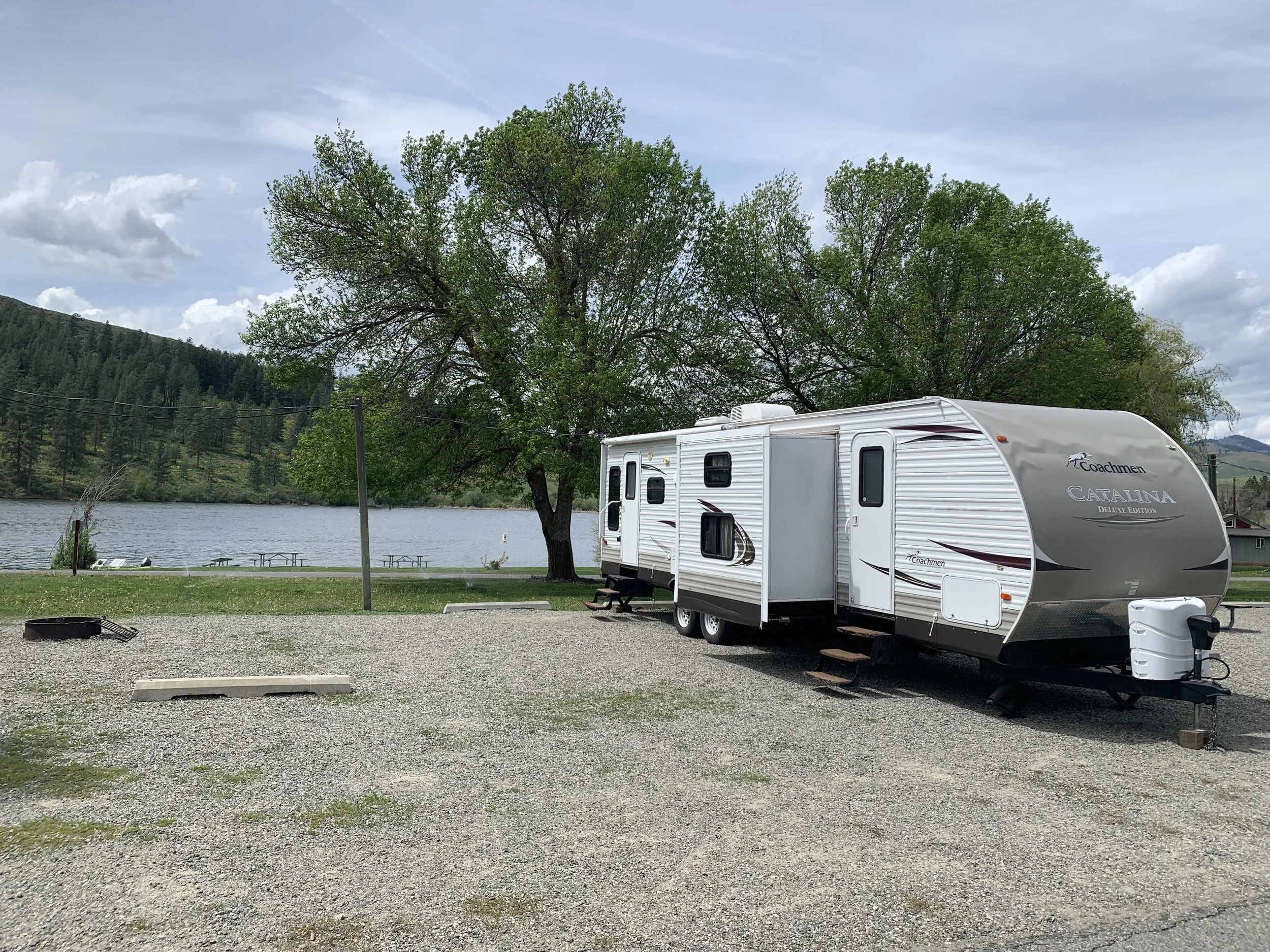 A recreational vehicle parked near a lake with trees and hills in the background, and picnic tables by the water.