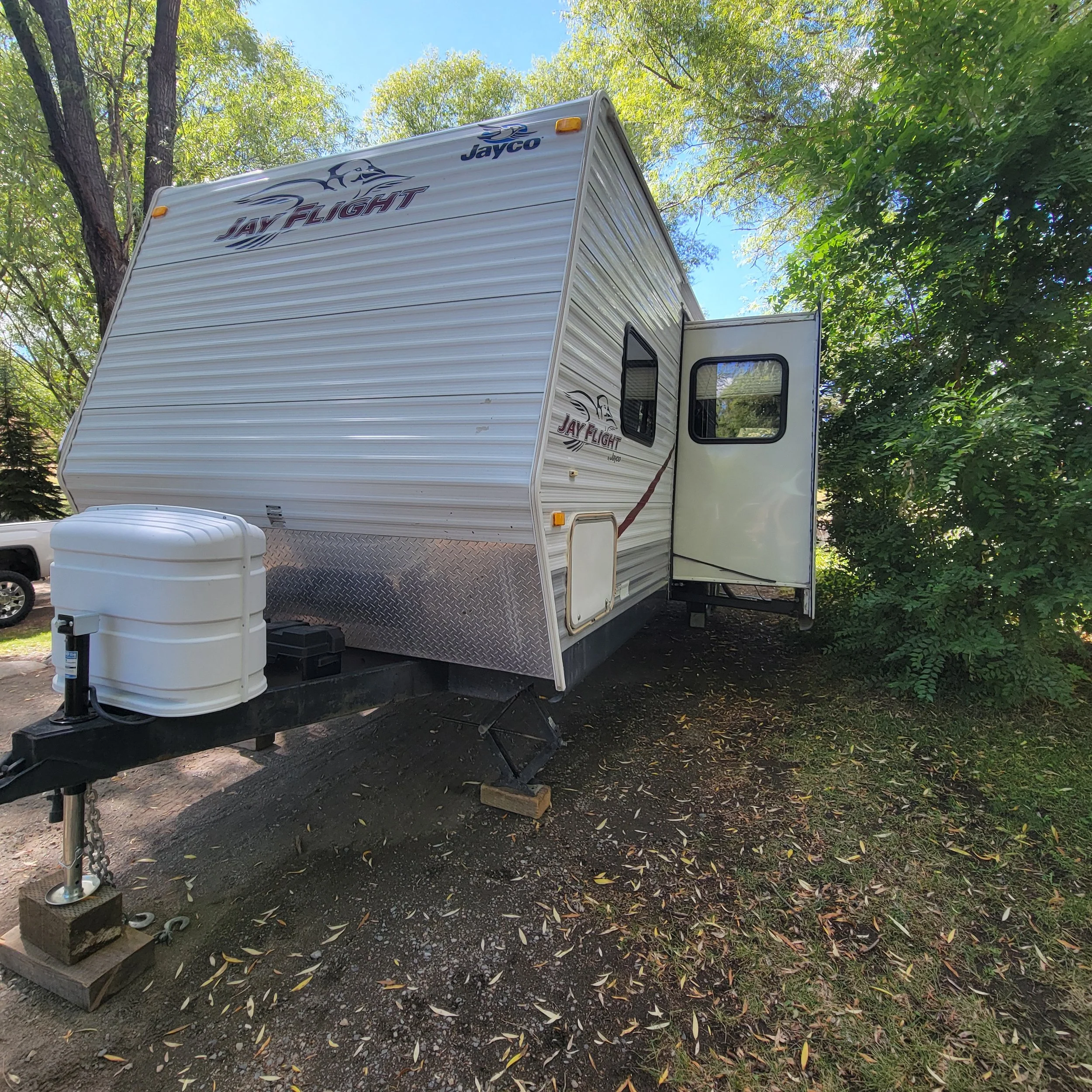 White Jayco Jay Flight travel trailer parked on a dirt surface surrounded by green trees under a blue sky.