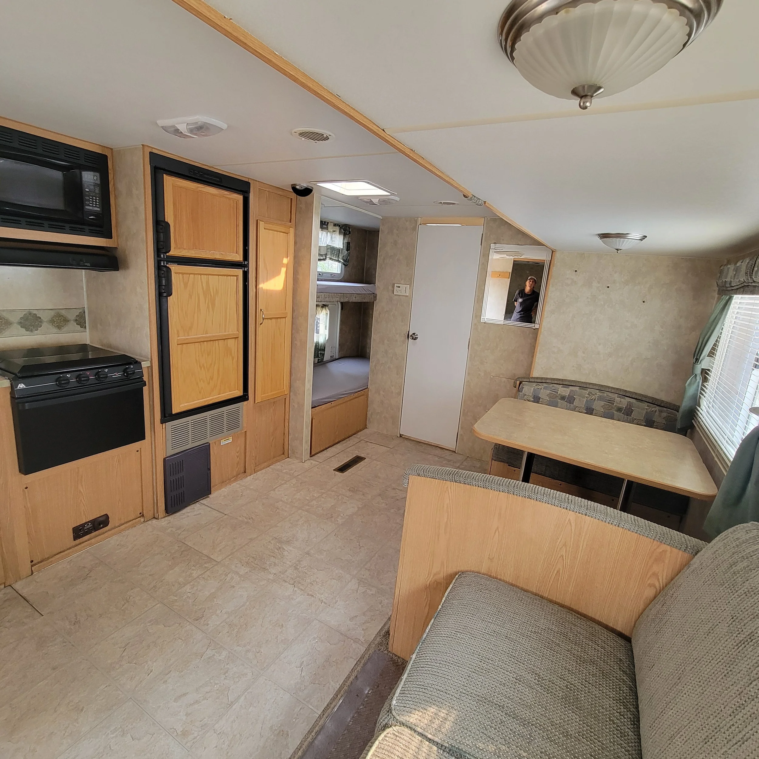 Interior of a small living space with a kitchenette on the left, including a microwave, stove, and refrigerator with wooden cabinet doors. In the middle, there are two built-in bunk beds with bedding. On the right, there is a dining area with a corne