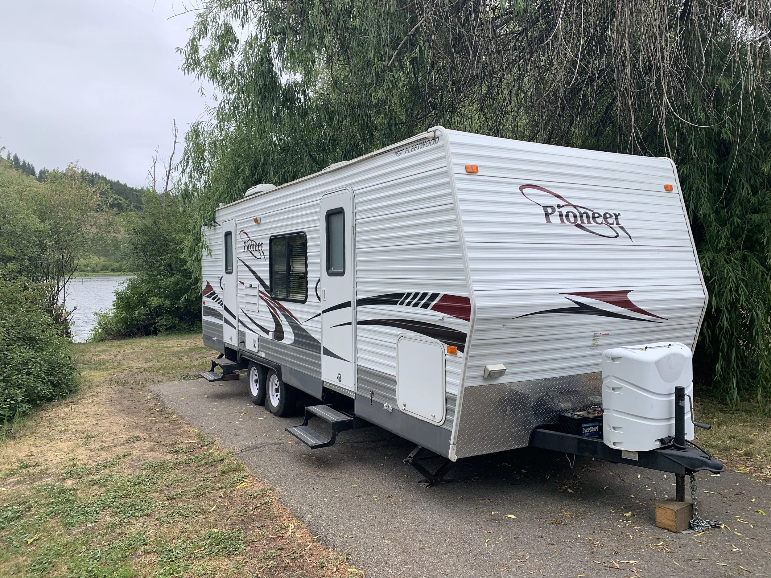 A white Pioneer travel trailer with red and black graphics parked on a paved site near a lake, surrounded by trees.