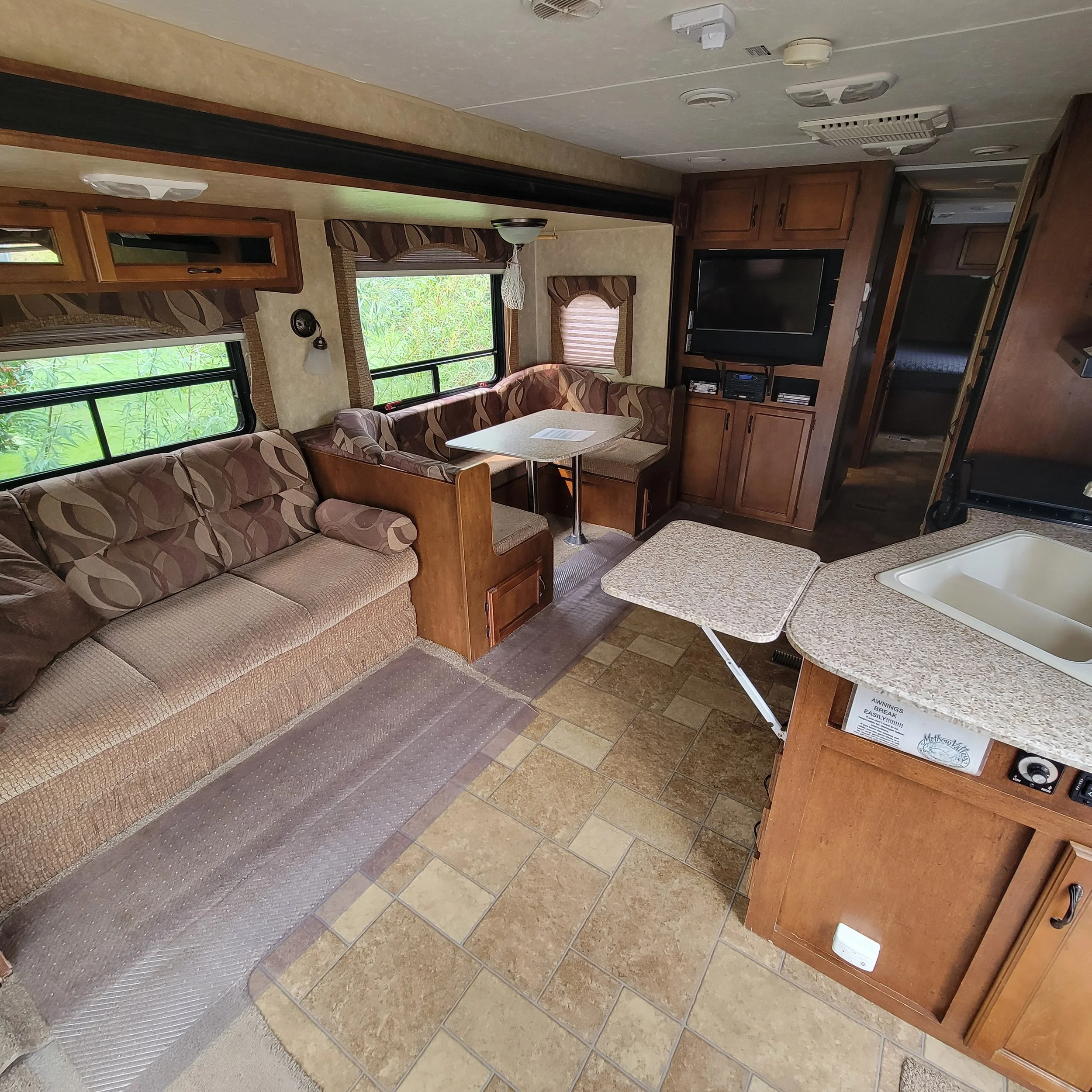 Interior of an RV with a beige couch, a dining nook with patterned chairs, a TV, wooden cabinets, and a kitchen with a sink and countertops.