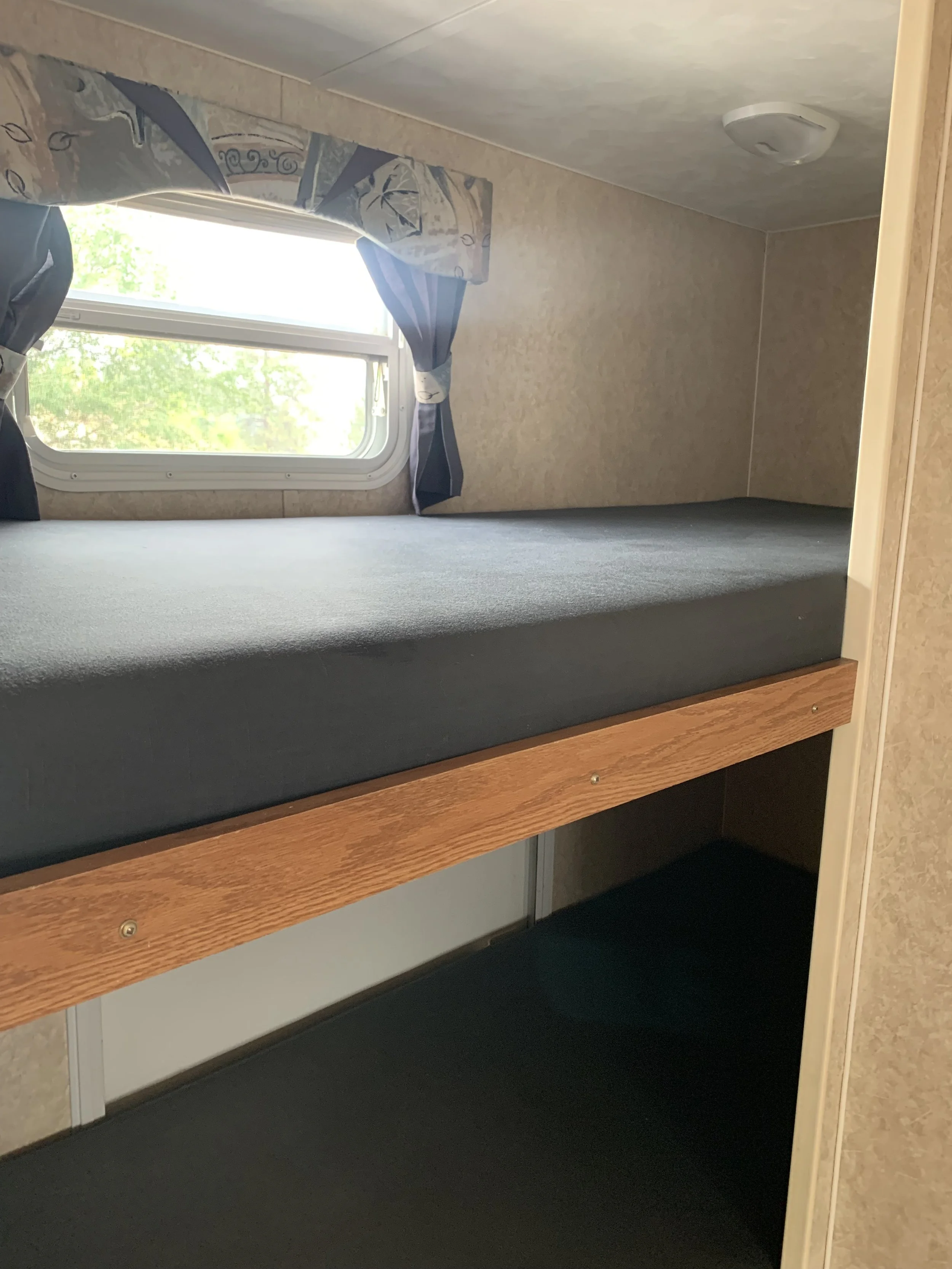 Interior view of a sleeping area inside a camper or RV with a bunk bed, small window with curtains, and beige walls.