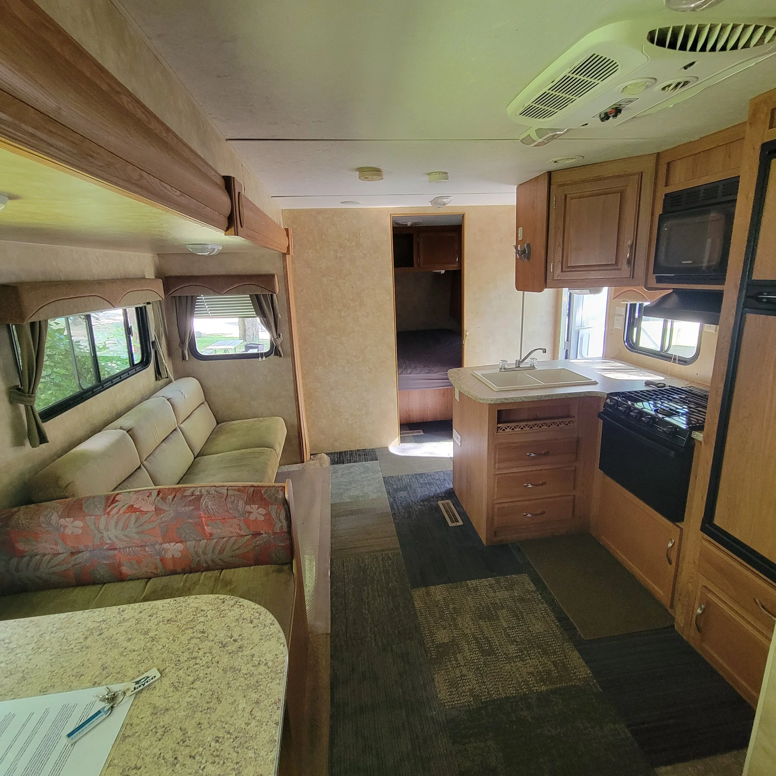 Interior of a camper with a kitchen area and living space, including a sofa, window with curtains, kitchen sink, stove, microwave, and cabinets.