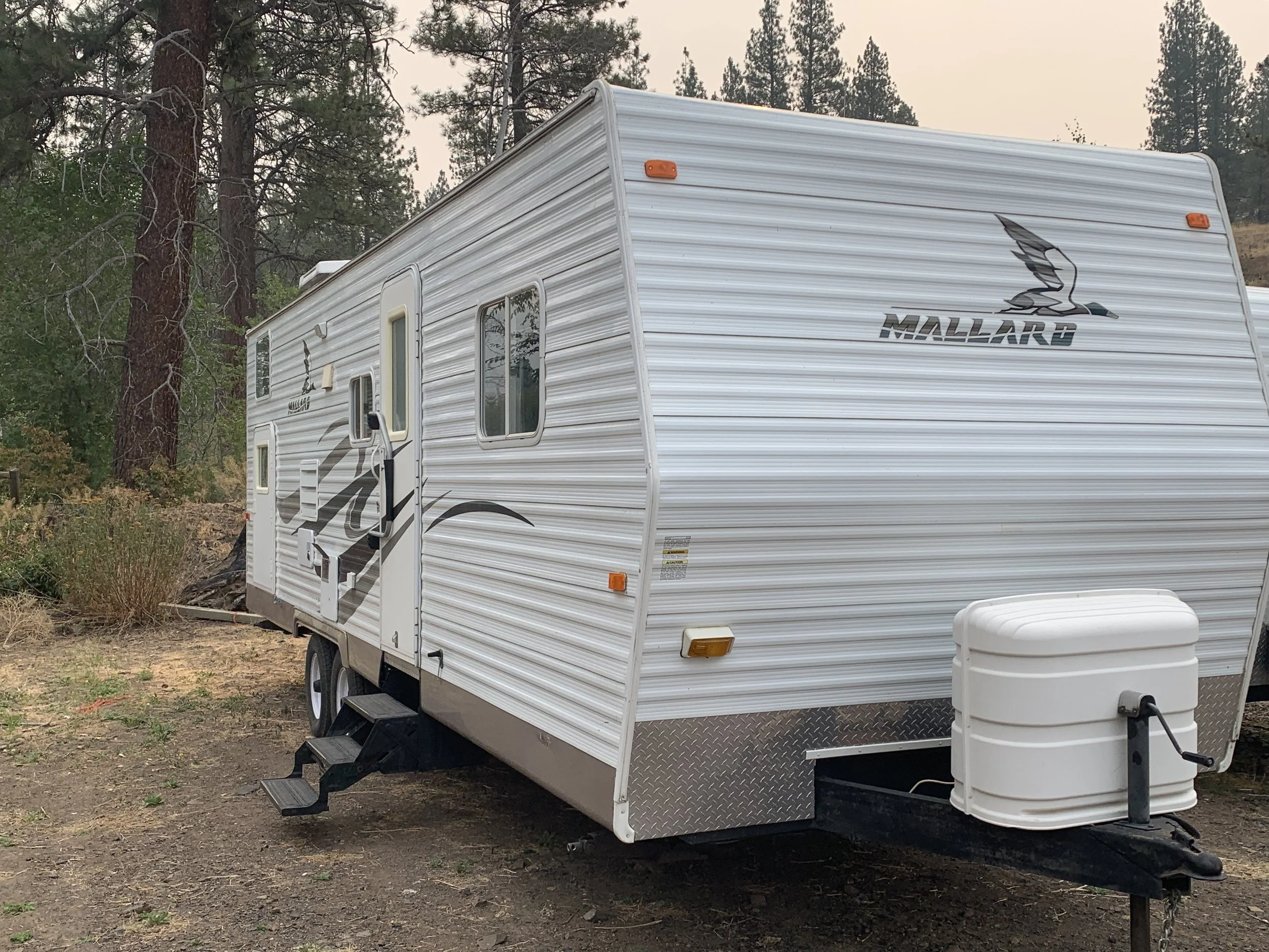 A white Mallard travel trailer with silver accents, parked on a dirt patch beside trees, with steps leading to the door and a propane tank attached at the front.