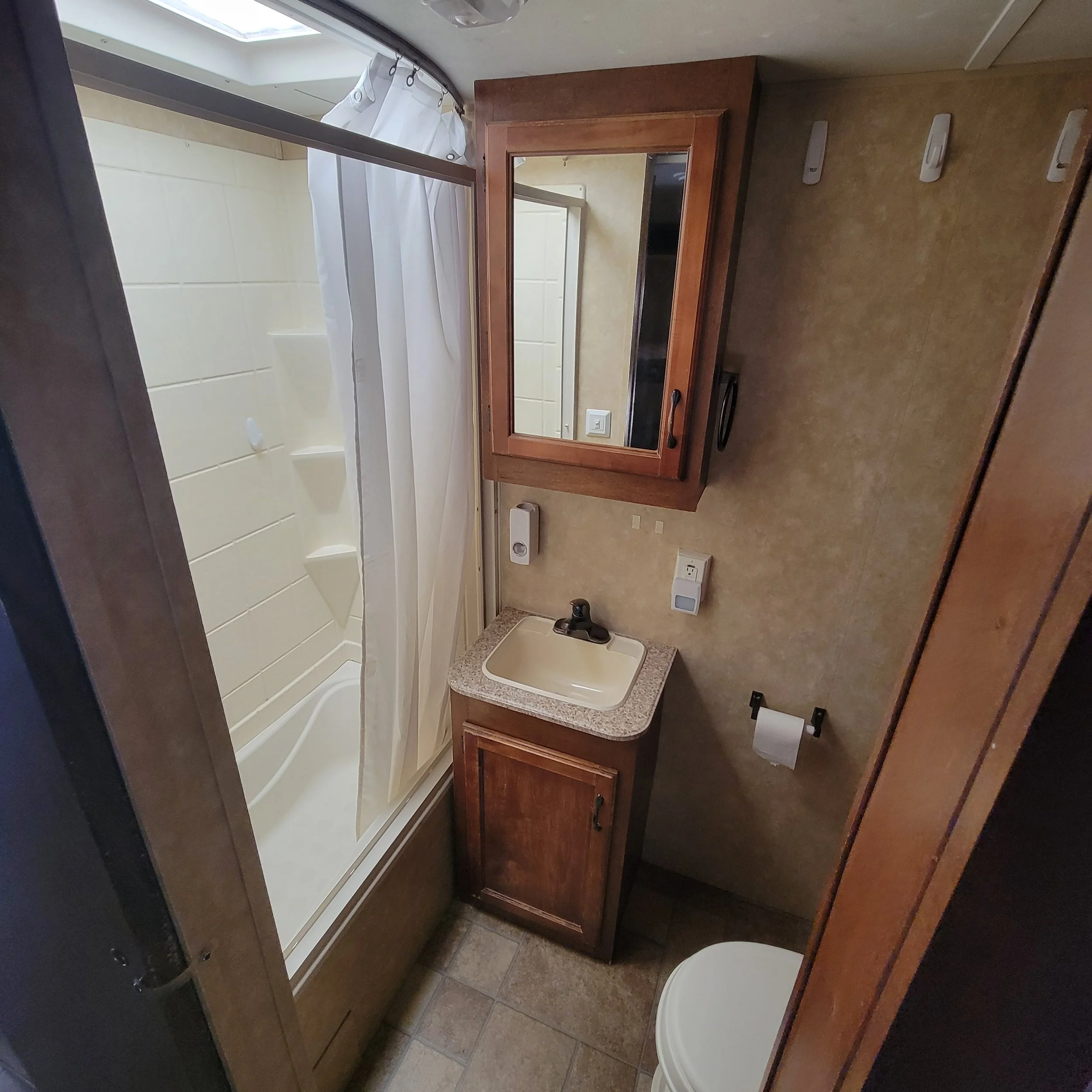 Small bathroom with a shower, sink, and mirror with a wooden cabinet. The shower has a sliding glass door and a white curtain. Toilet paper holder next to the toilet. Tiled floor and beige walls.
