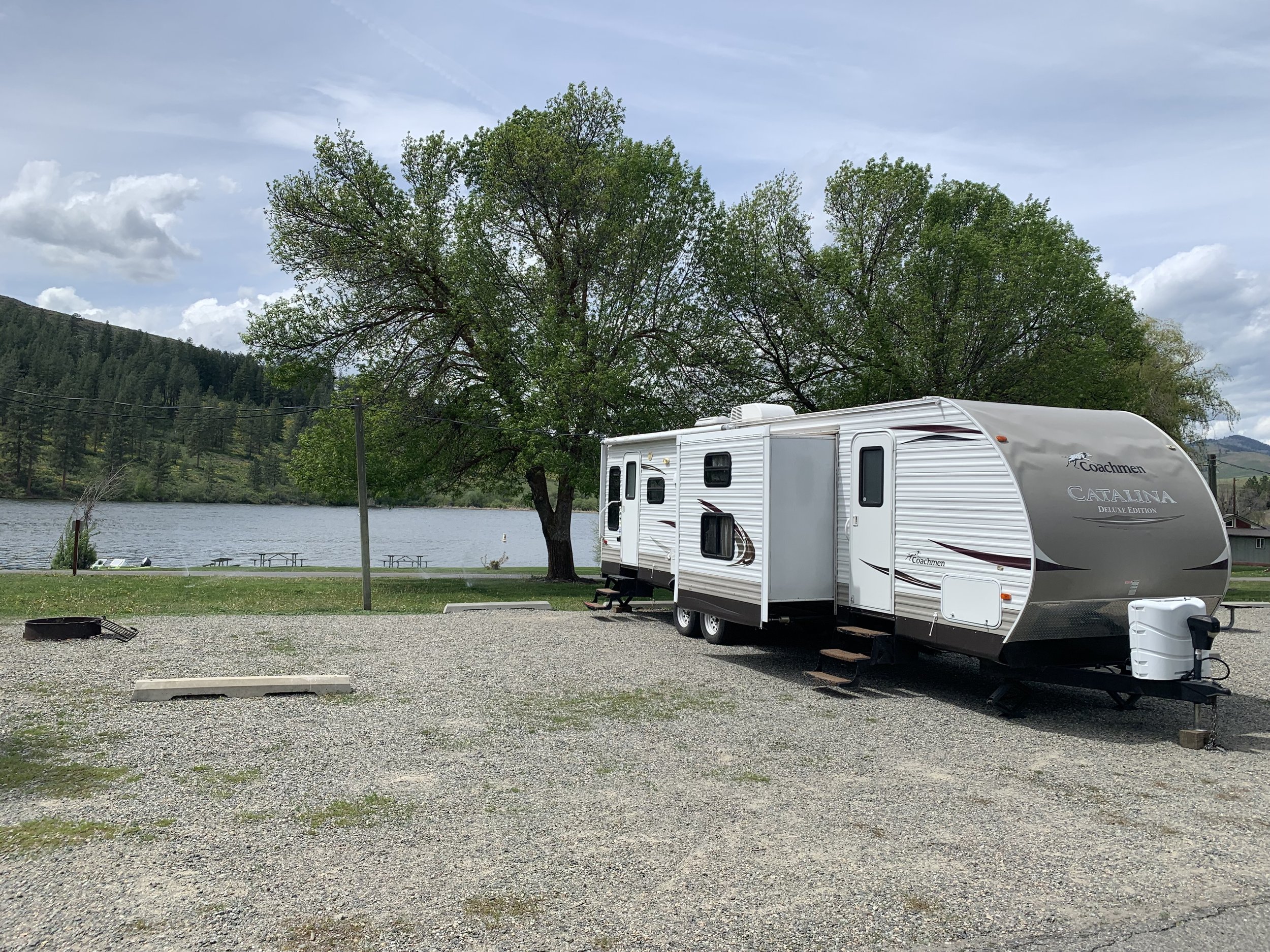 A camping trailer parked on a gravel lot near a lake with trees and mountains in the background.